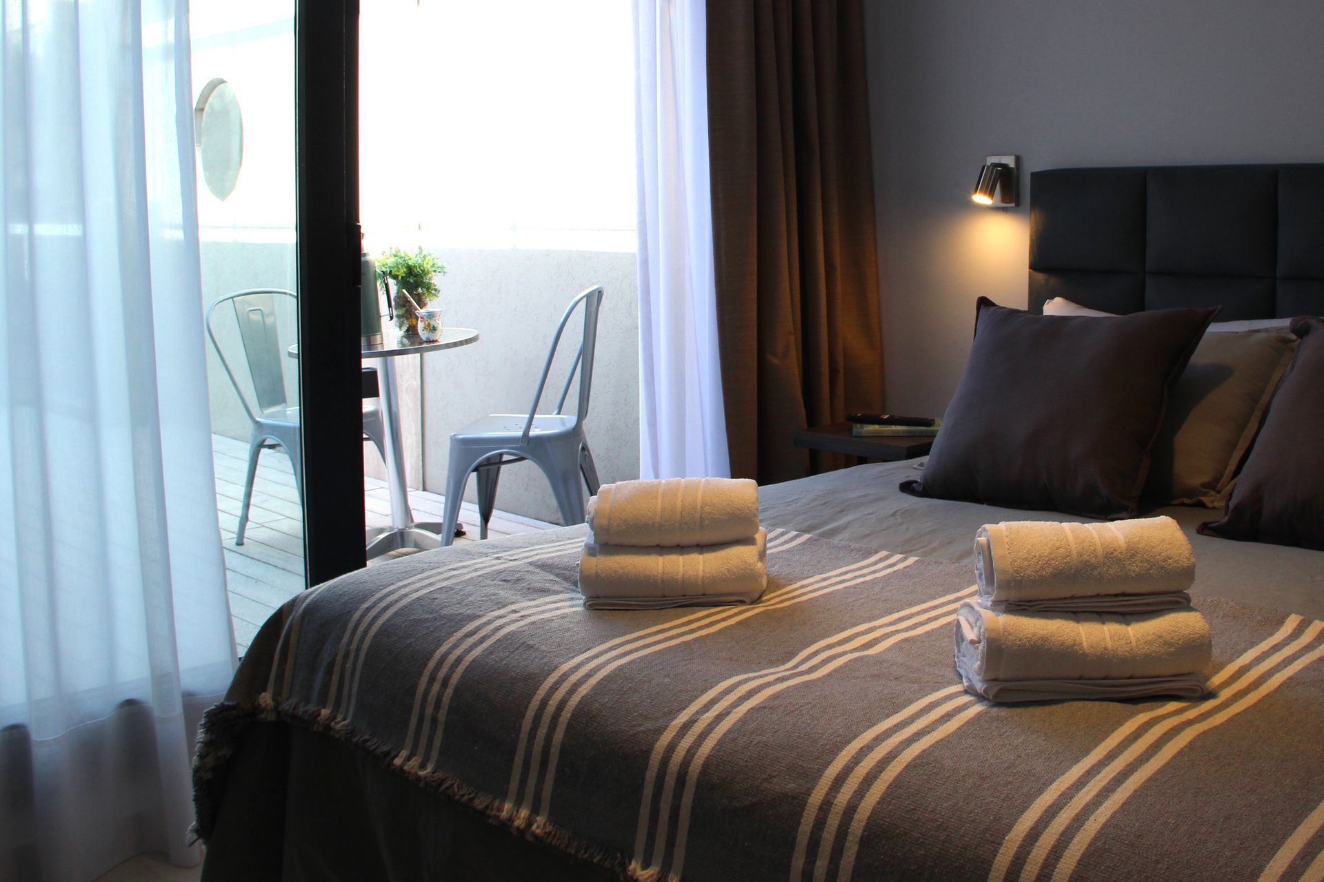 Modern hotel room with neatly made bed, folded towels, and balcony seating by a window