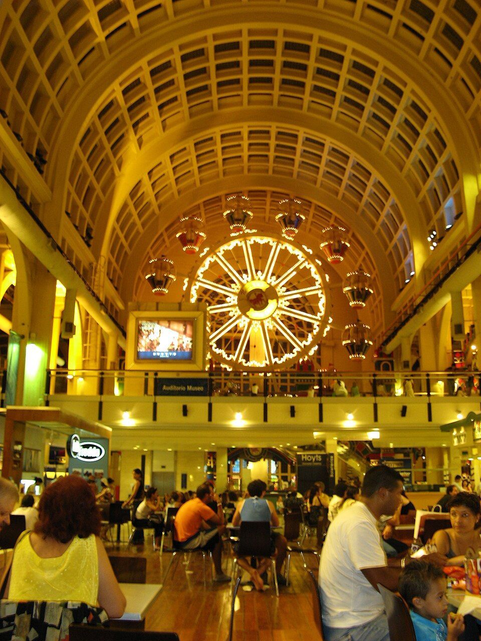Grand hall with a large illuminated clock, arched ceiling, and people seated at tables below