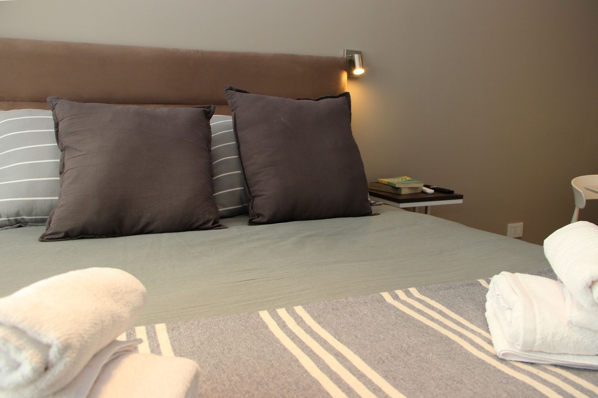 Neatly made bed with gray striped bedding, two dark pillows, towels, and a bedside lamp in a beige bedroom