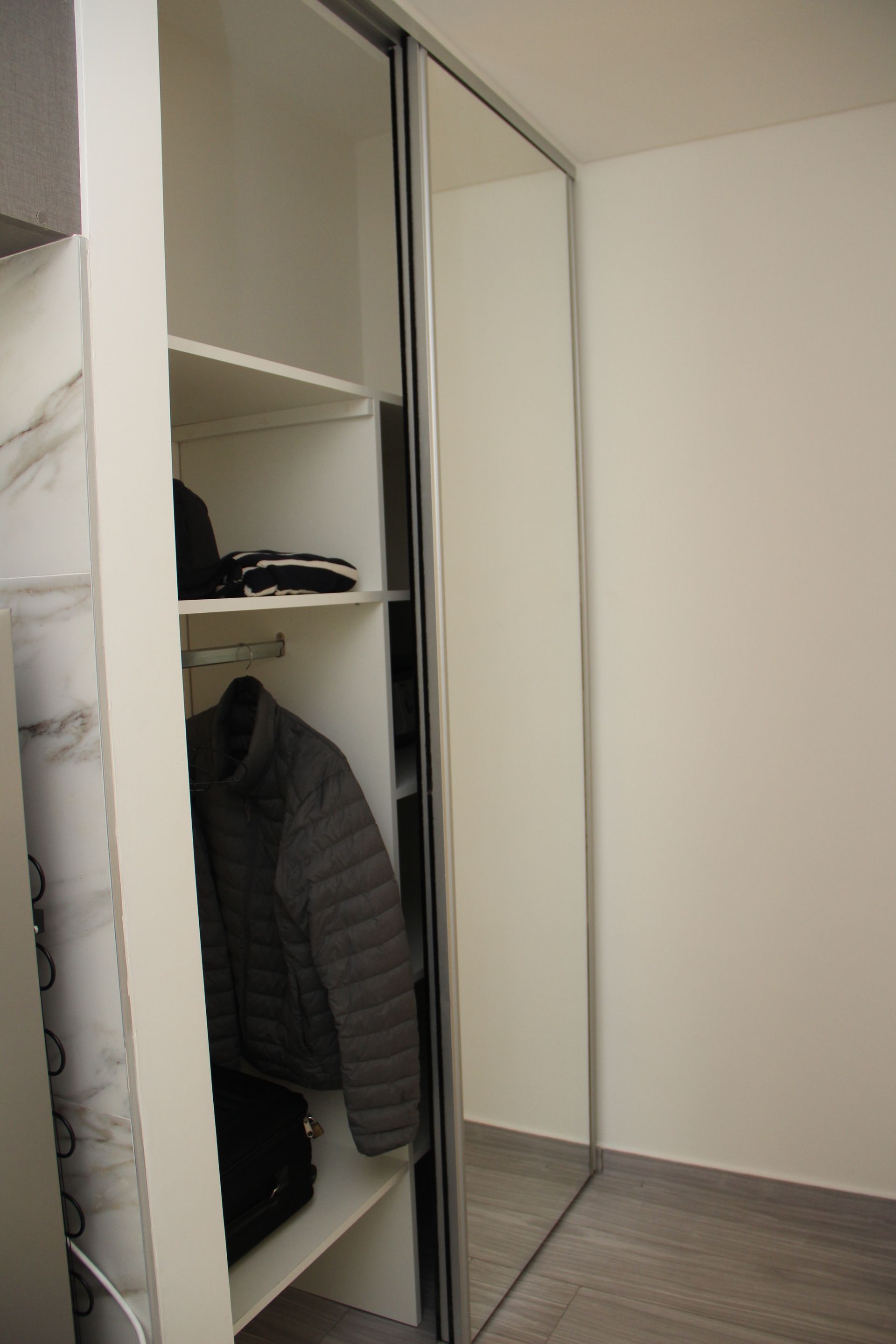 Open closet with mirrored sliding doors, shelves, hanging jackets, and a gray floor in a bare room