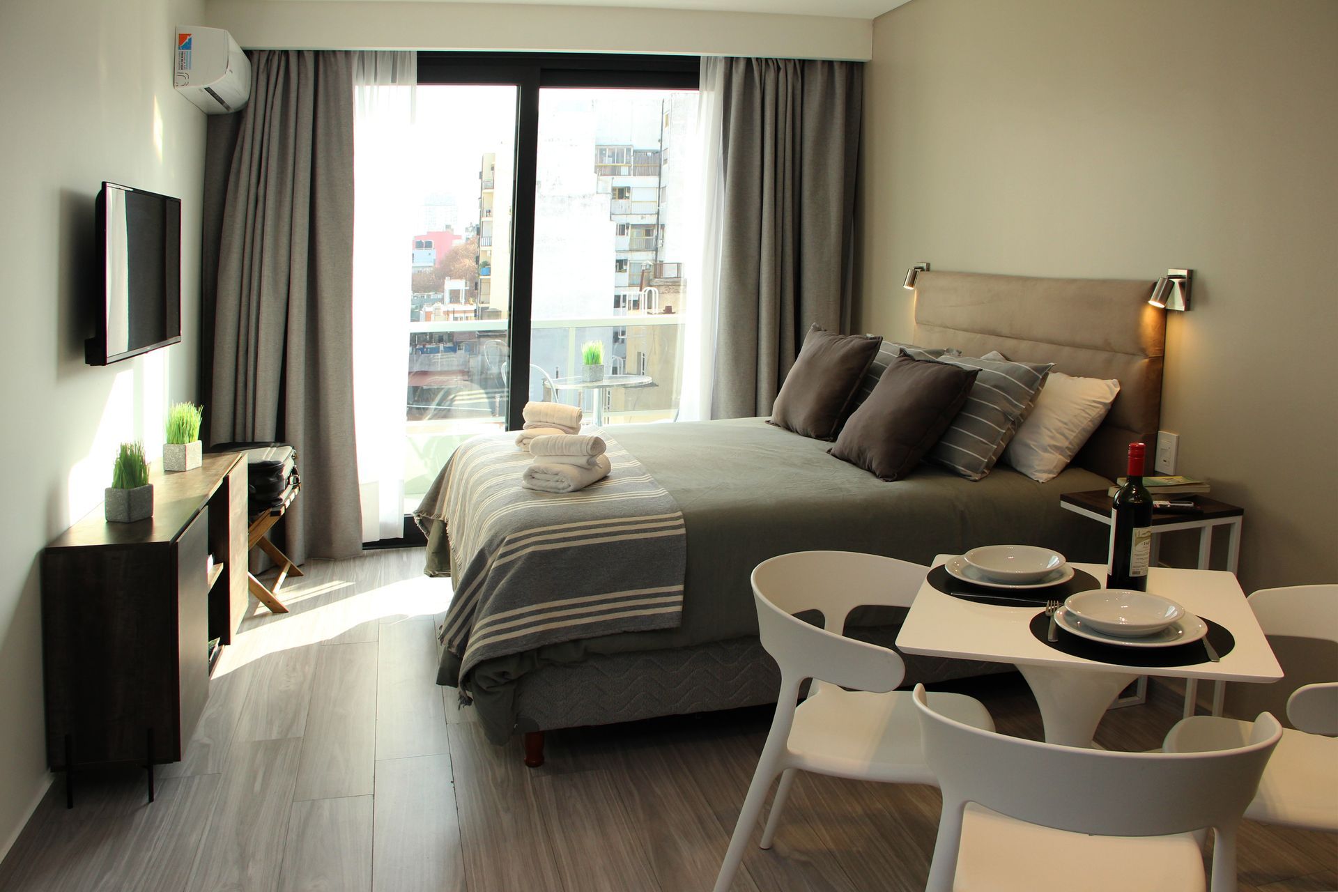 Modern studio bedroom with a neatly made bed, small dining table, and balcony doors letting in daylight
