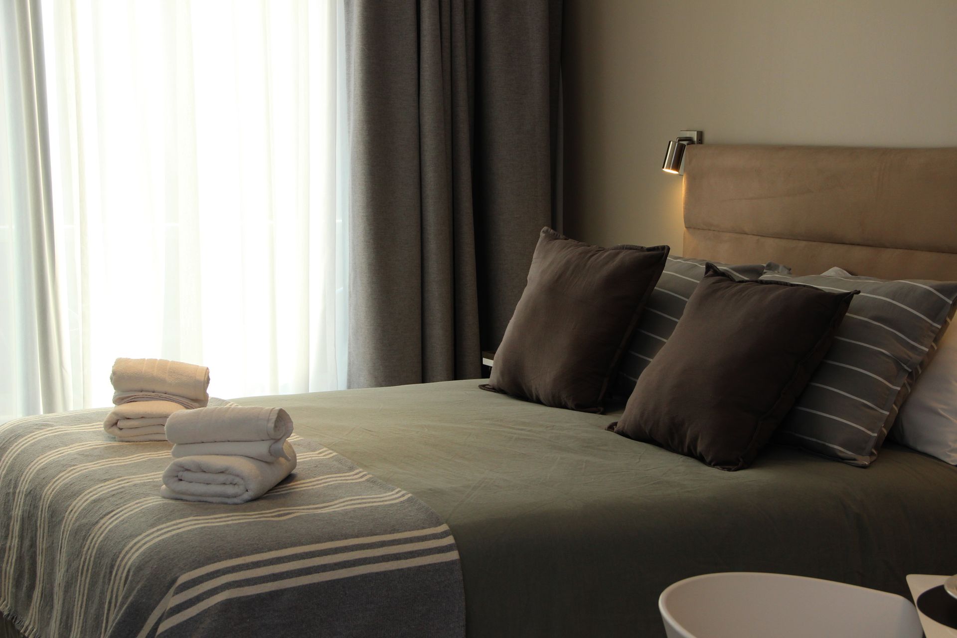 Neatly made bed with gray bedding, pillows, folded towels, and curtains by a sunlit window.