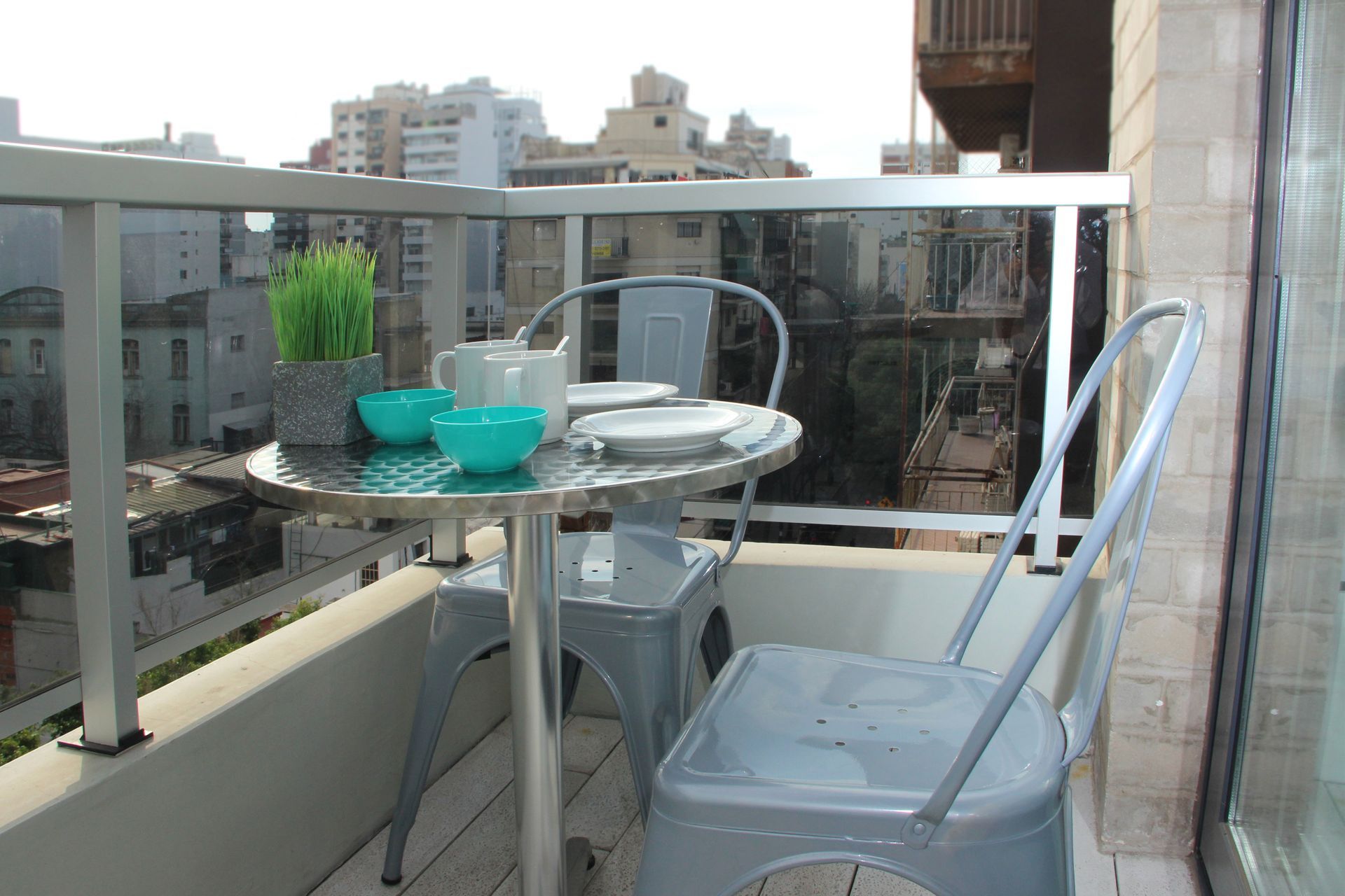 Balcony with glass railing, metal table and chairs, and turquoise bowls overlooking city buildings