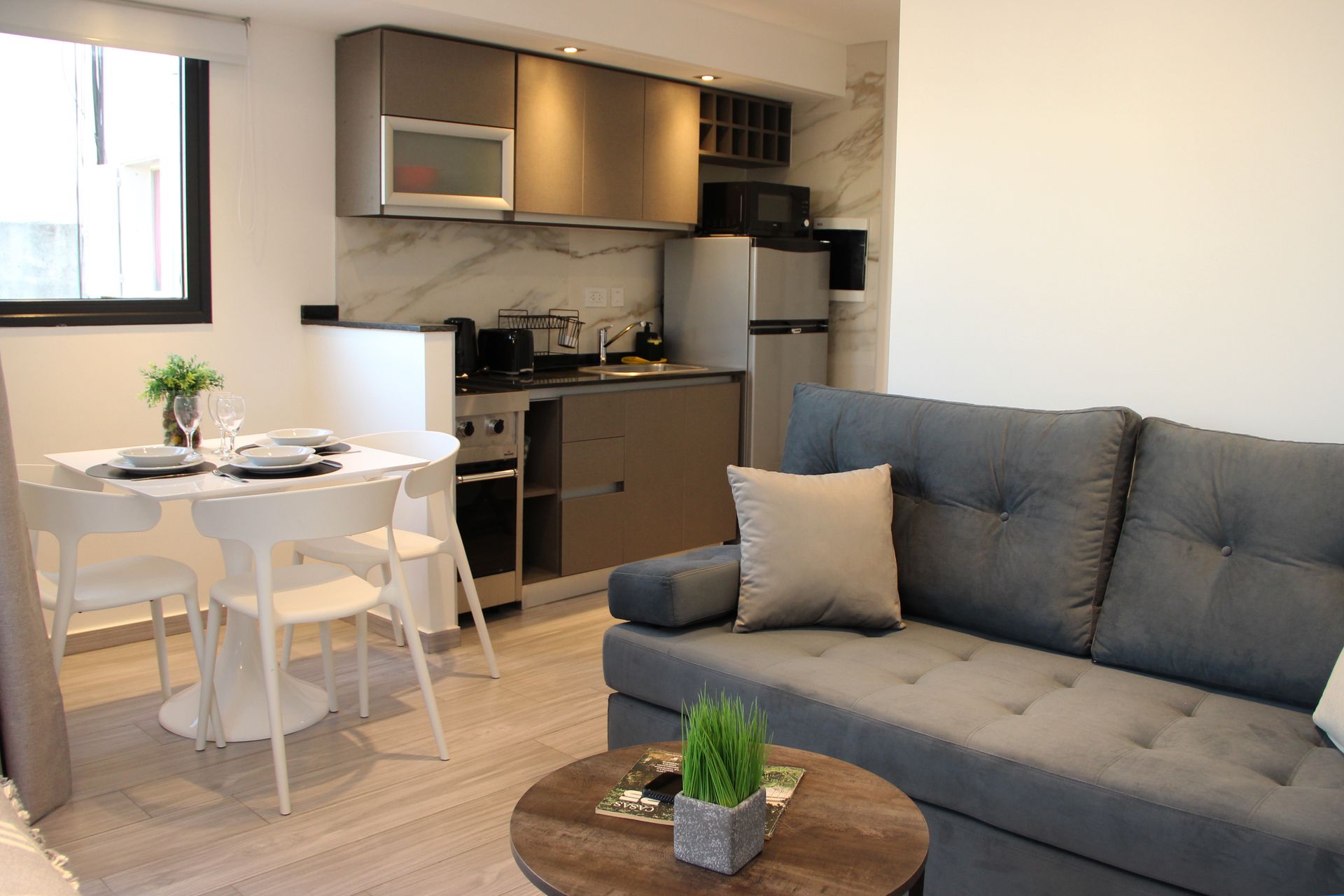 Modern apartment living room with gray sofa, small dining table, and kitchenette in the background
