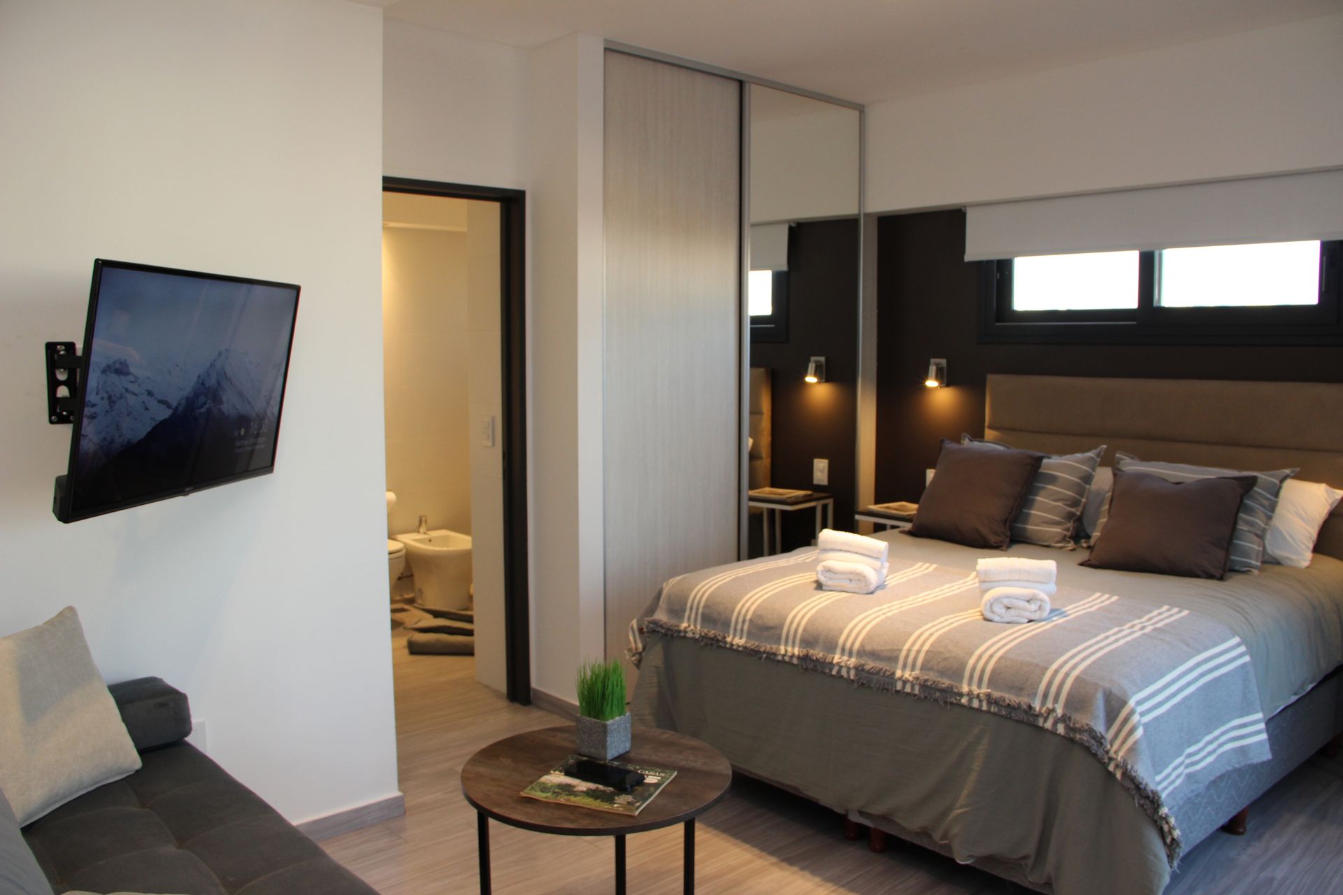 Modern bedroom with a neatly made bed, wall-mounted TV, and a small round table in the foreground