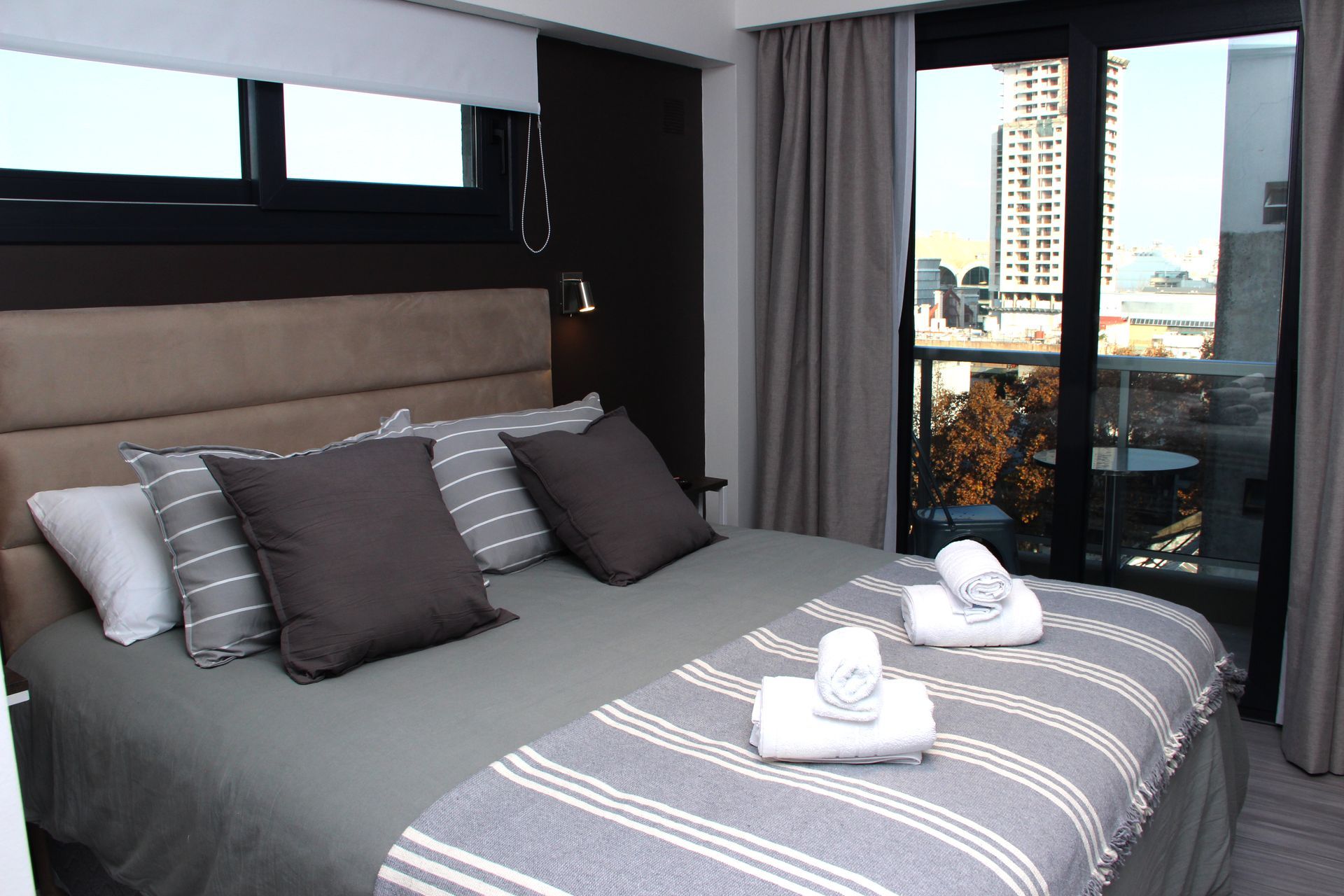 Modern bedroom with a bed made up in gray and white linens, overlooking a city view through large windows.