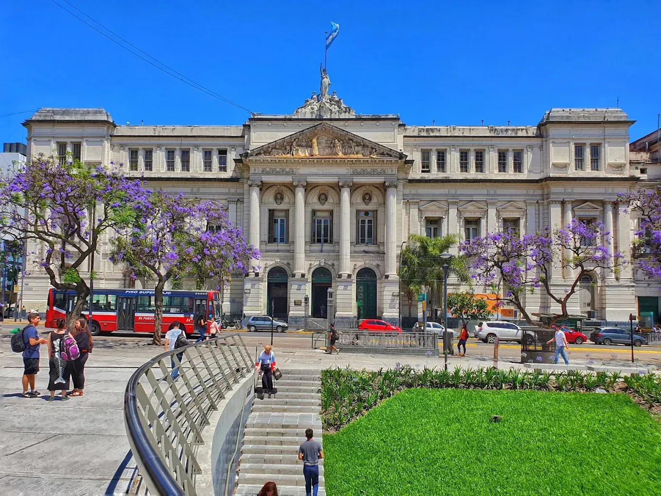 Historic civic building with grand columns, people in plaza, purple trees, and a green lawn under blue sky