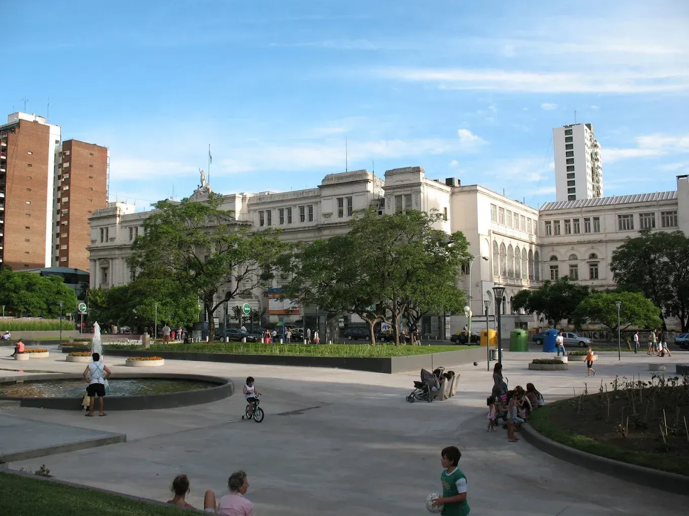 City park with people walking and biking beside a fountain, framed by historic buildings under a blue sky
