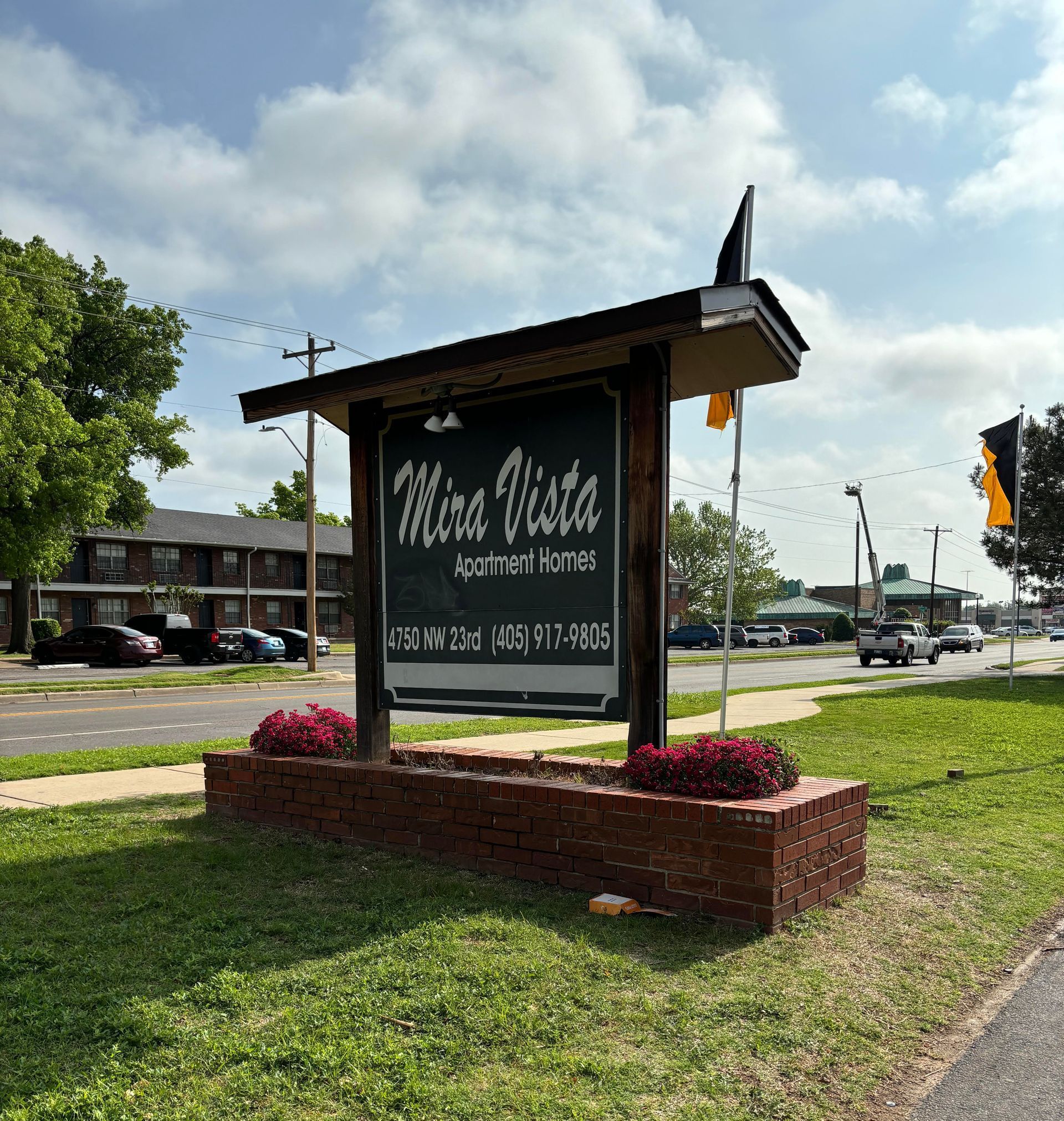Mira Vista Apartment Homes sign on a brick base with flowers, road in front, apartments in the background.