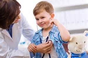 Doctor examining a smiling young boy's chest with a stethoscope in a medical office; a stuffed bear is nearby.