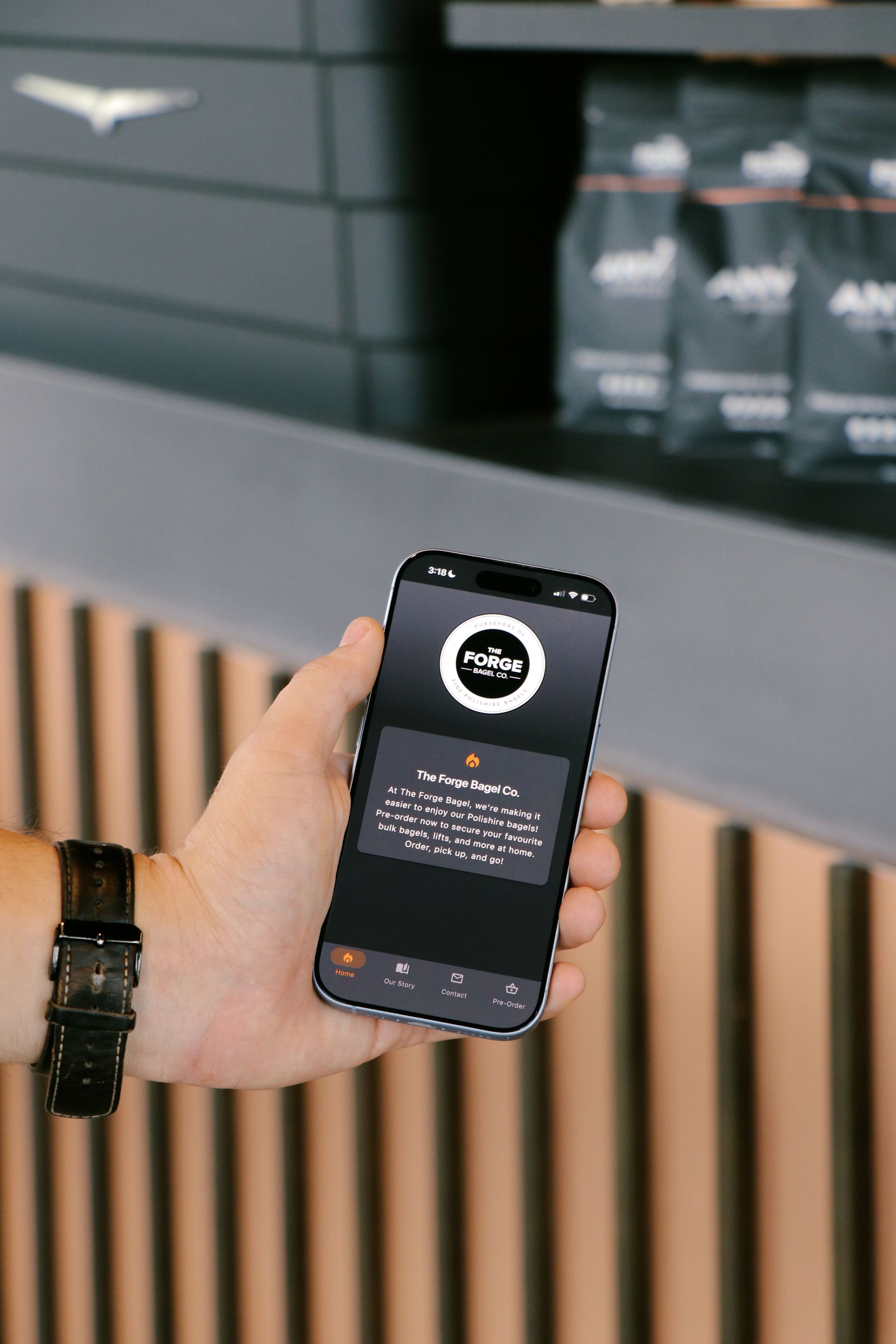 Hand holding a smartphone showing a dark app screen in a café-like setting with wood slats and shelves behind.