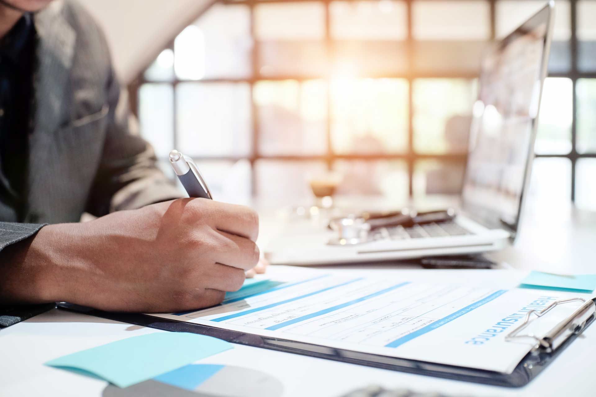 Person writing on a document at a desk with a laptop and bright window in background. Person writing on a document at a desk with a laptop and bright window in background.