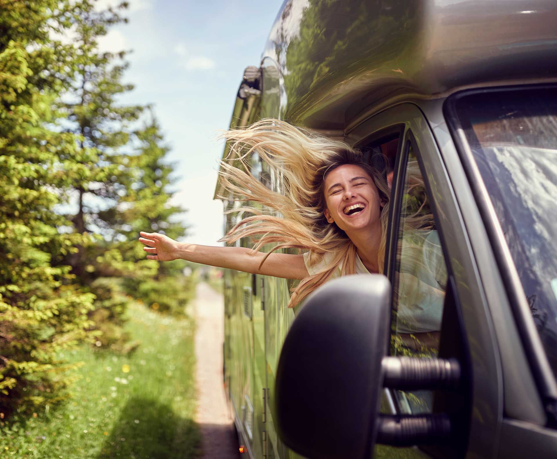Woman with blonde hair and outstretched arm, smiles from the window of a moving camper van, scenic roadside view. Woman with blonde hair and outstretched arm, smiles from the window of a moving camper van, scenic roadside view.