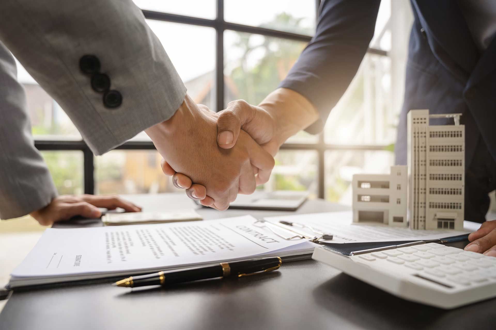 Two people shaking hands over a contract on a desk, with a model building and calculator nearby. Two people shaking hands over a contract on a desk, with a model building and calculator nearby.