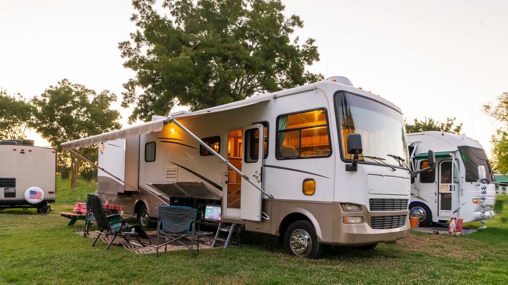RV parked in grassy area, awning extended, lit interior, camping chairs, small RV behind.