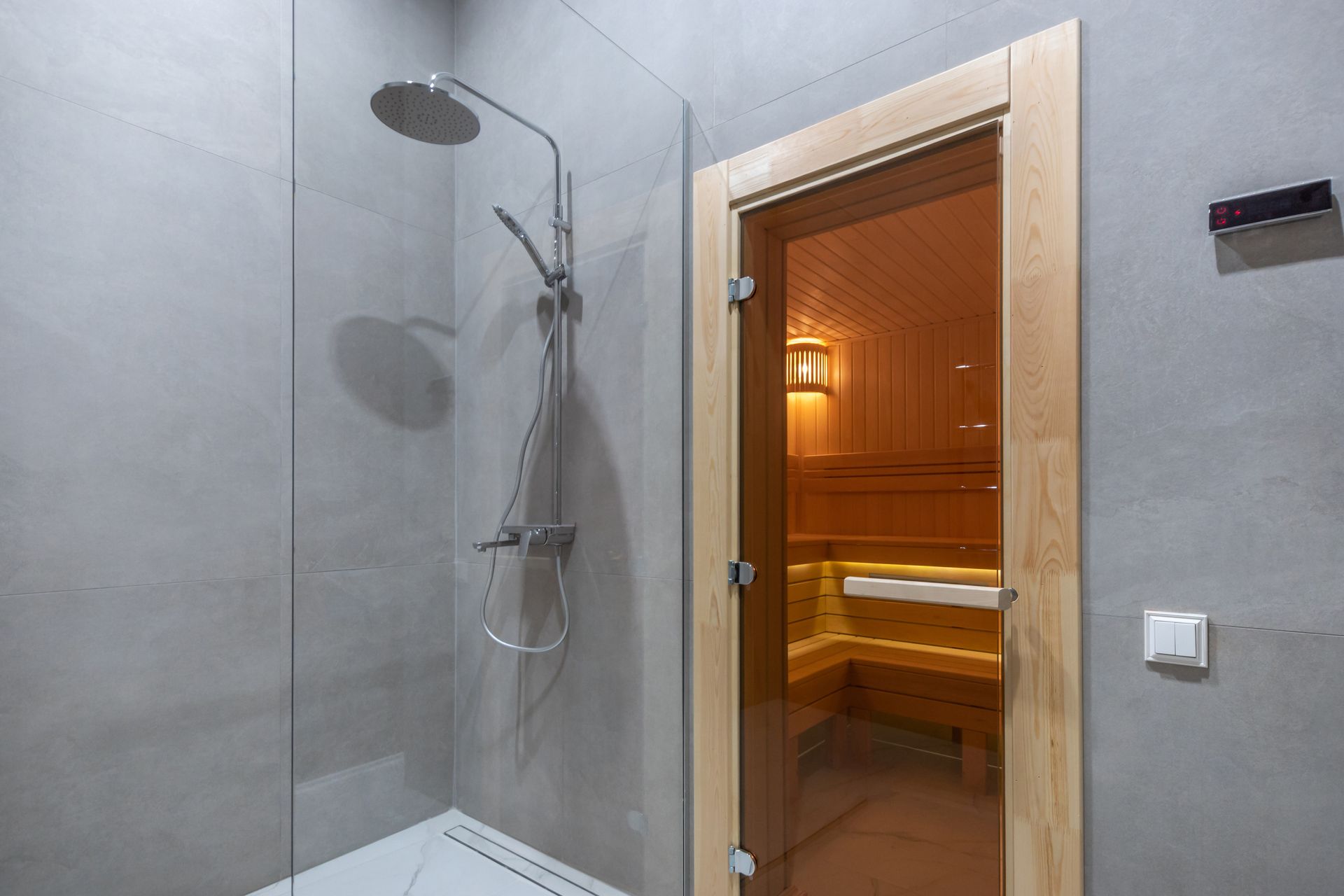 A modern shower with a glass partition and rainfall head adjacent to a wooden sauna with a glass door.