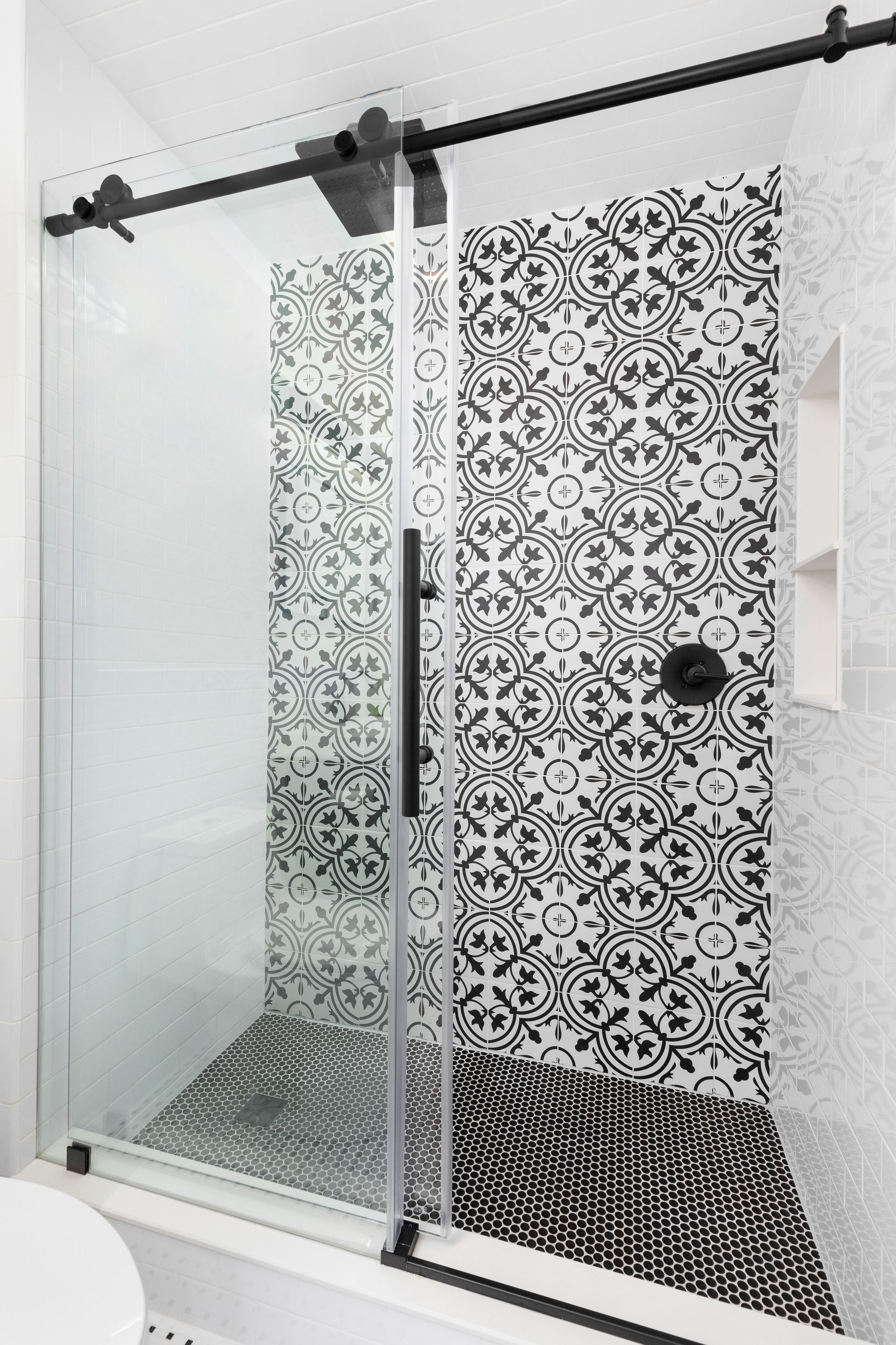 A walk-in shower with a black sliding glass door, patterned black and white wall tiles, and a dark pebble floor.