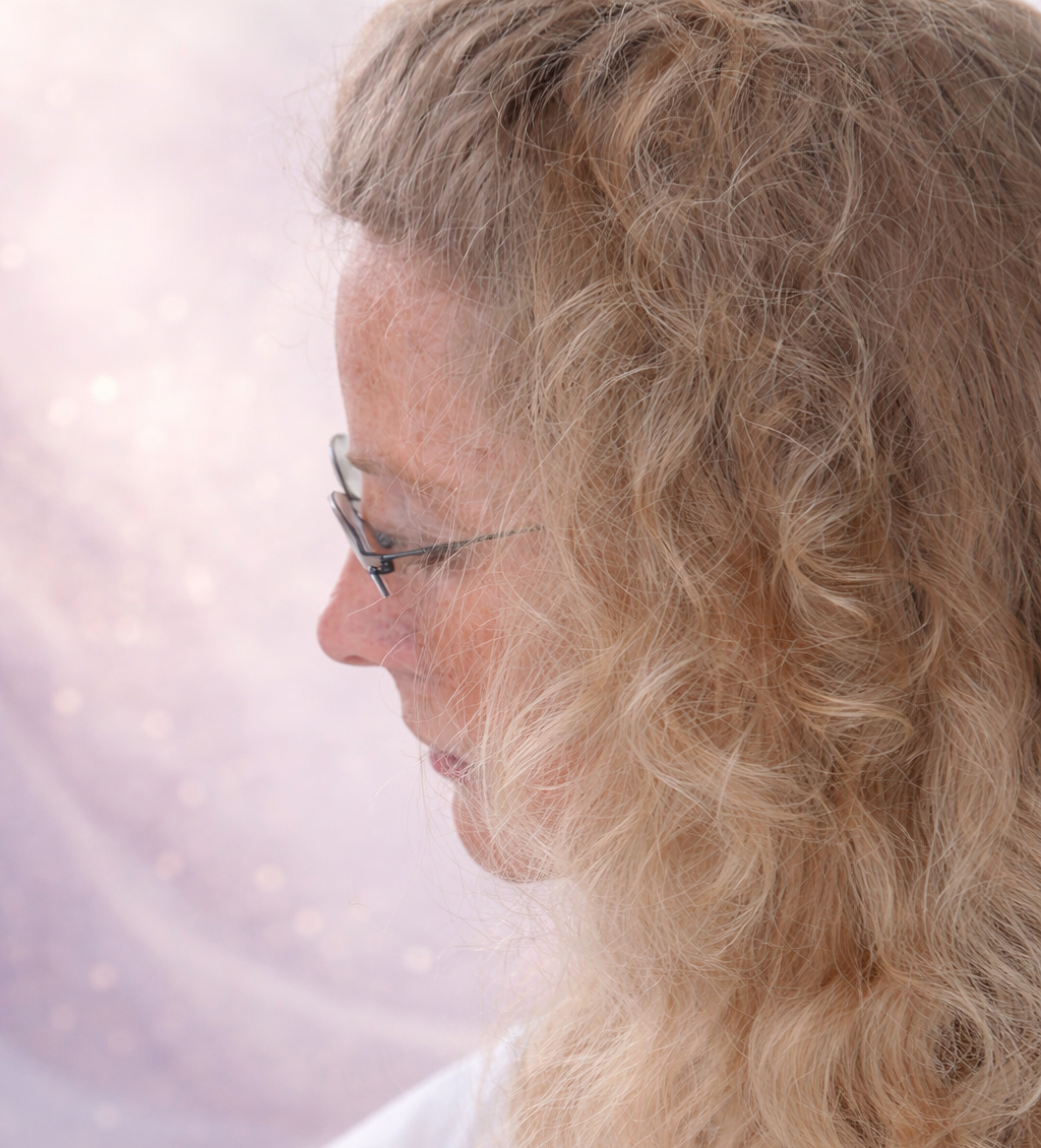 Anita met krullend blond haar en een bril, naar beneden kijkend, afgebeeld tegen een zachtroze achtergrond.