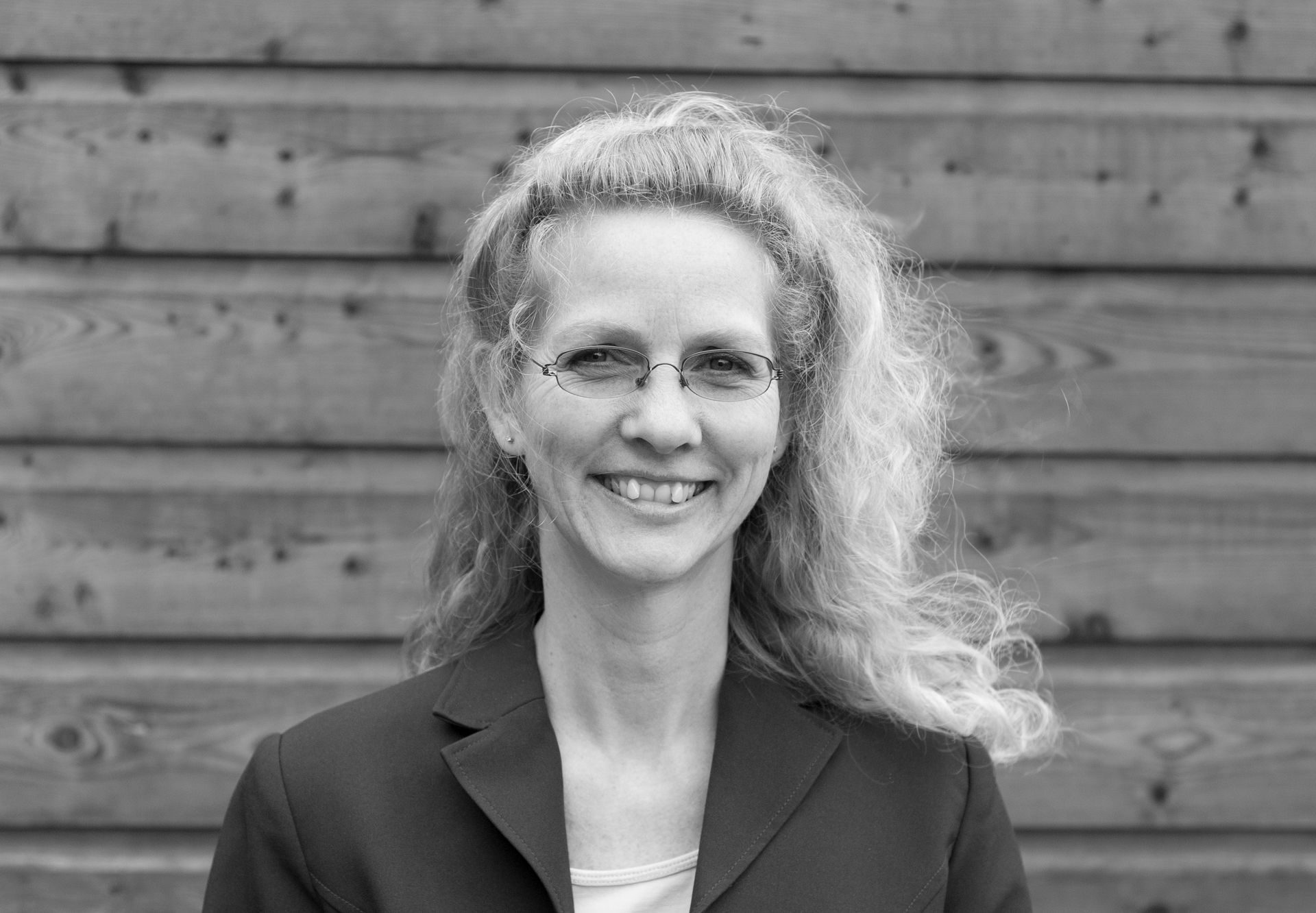 Anita, lachend, in een blazer, voor een houten lambrisering.