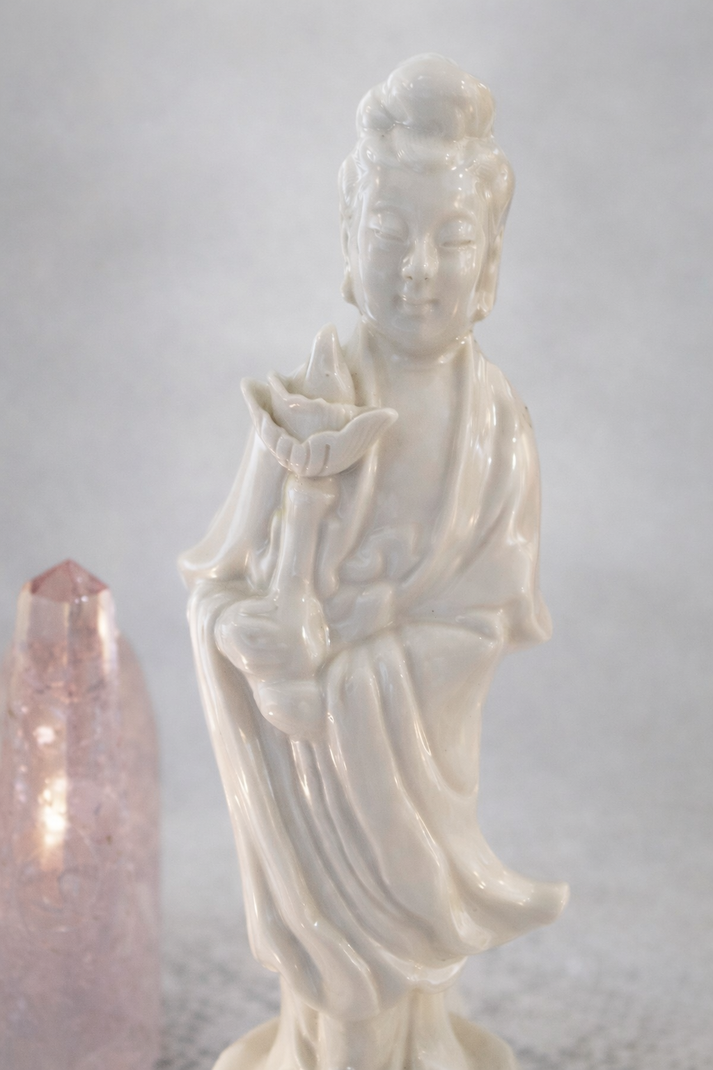 Een wit porseleinen beeldje van Guanyin die een lotusbloem vasthoudt, met een roze kristal op de achtergrond.