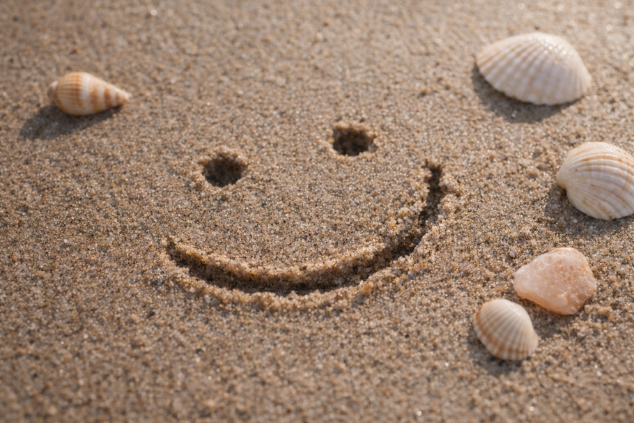 Een smileygezichtje getekend in het zand, met schelpen eromheen verspreid.