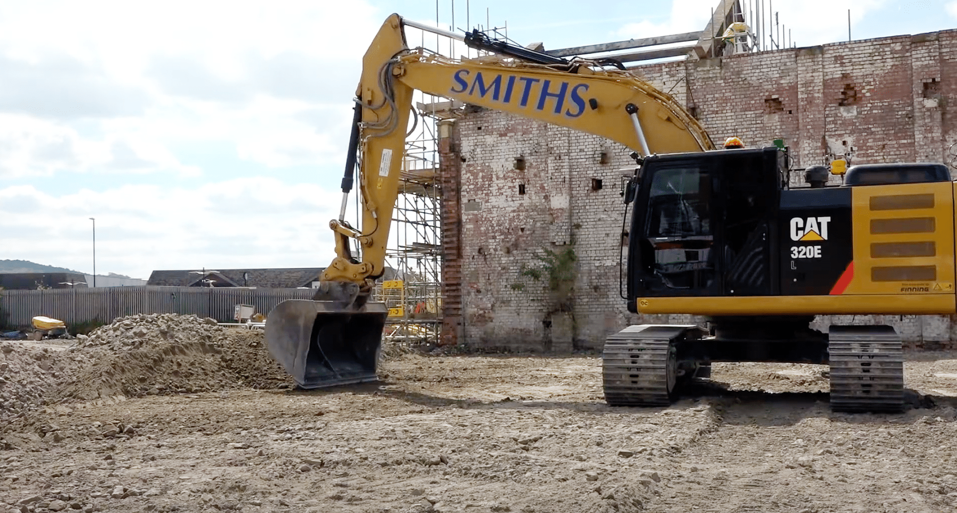 A large yellow excavator is sitting in a dirt field in front of a brick building.