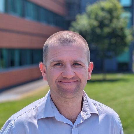 A man in a blue shirt is smiling in front of a building