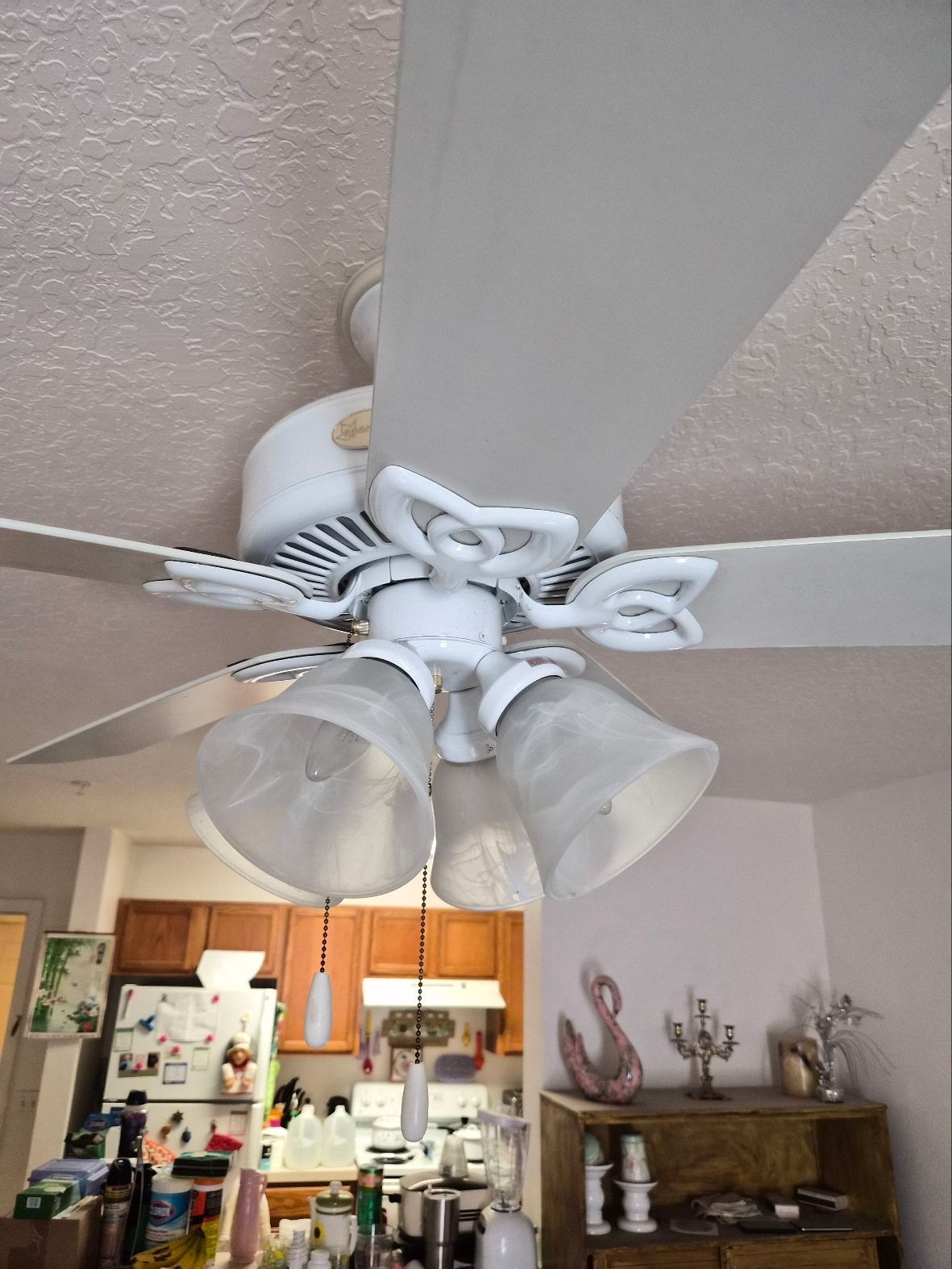 White ceiling fan with four frosted lights in a living room above a kitchen and bookshelf.