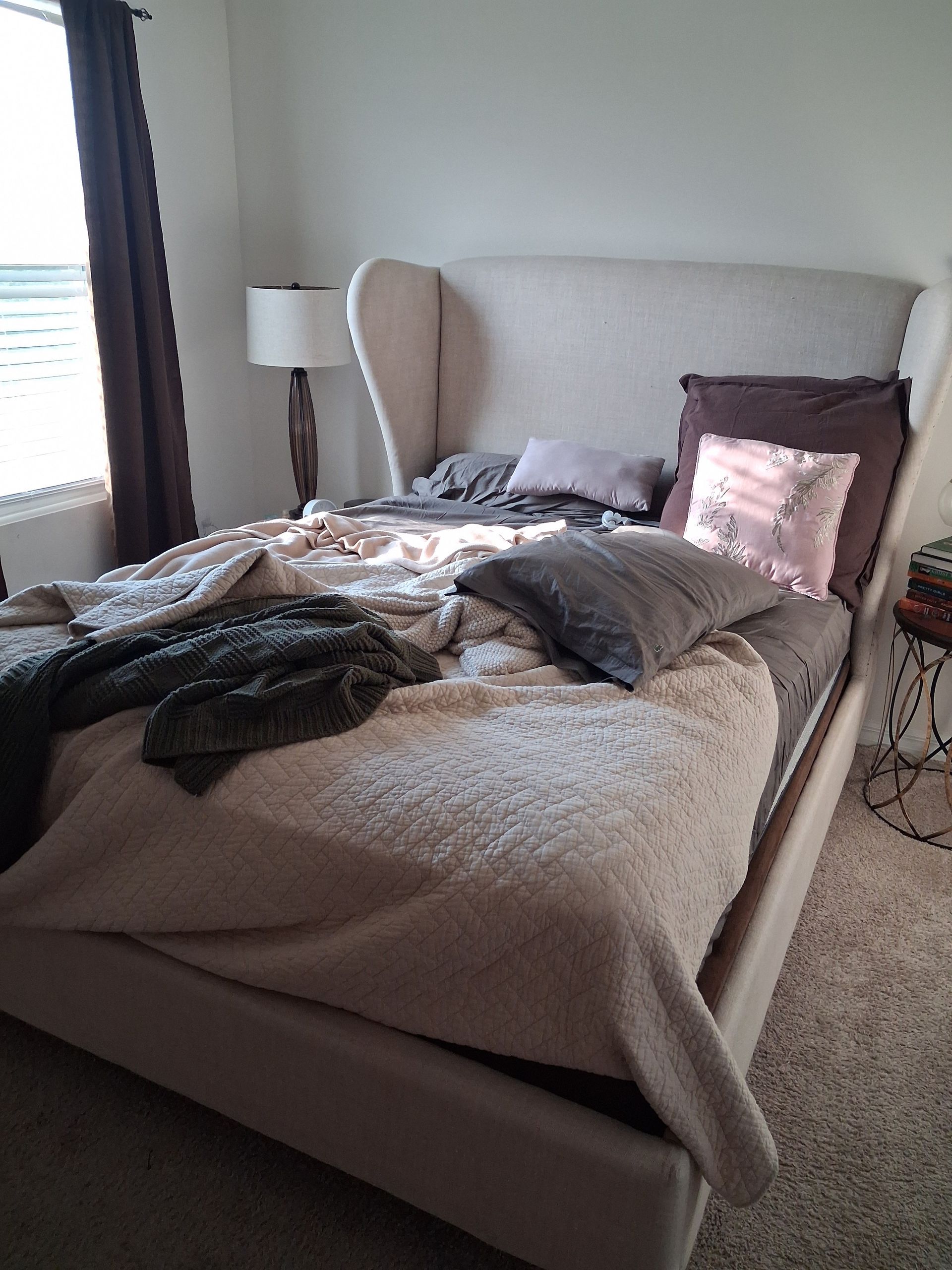 Unmade beige bed with pillows, blanket, and clothes in a softly lit bedroom near a window and lamp