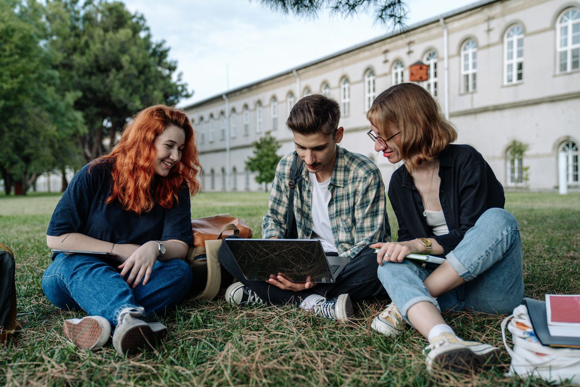 Three students seated on grass, looking at a laptop. One with red hair smiles. A large building is in the background.