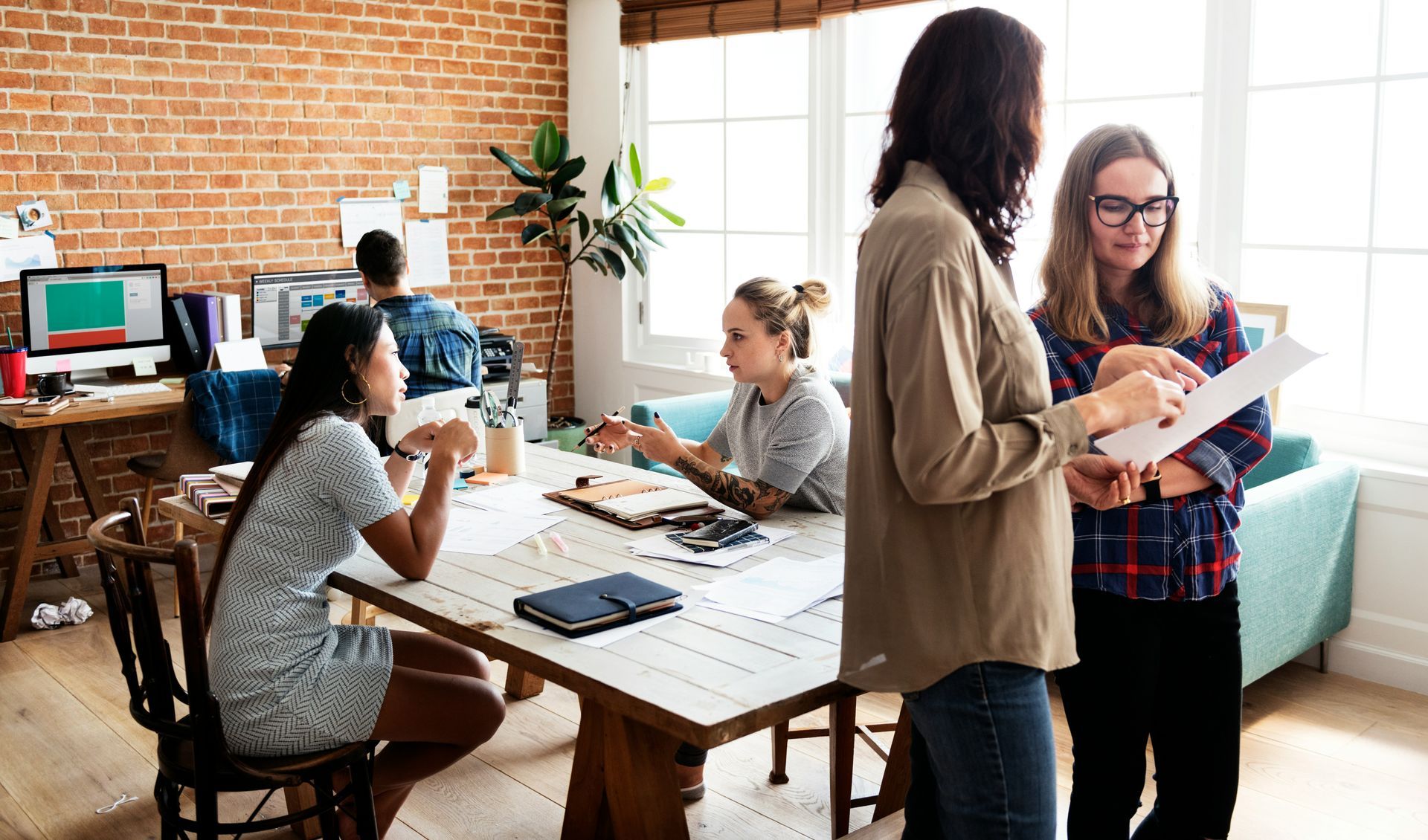 People working in a bright, brick-walled office. Two women discuss paperwork, others work at desks and a table.