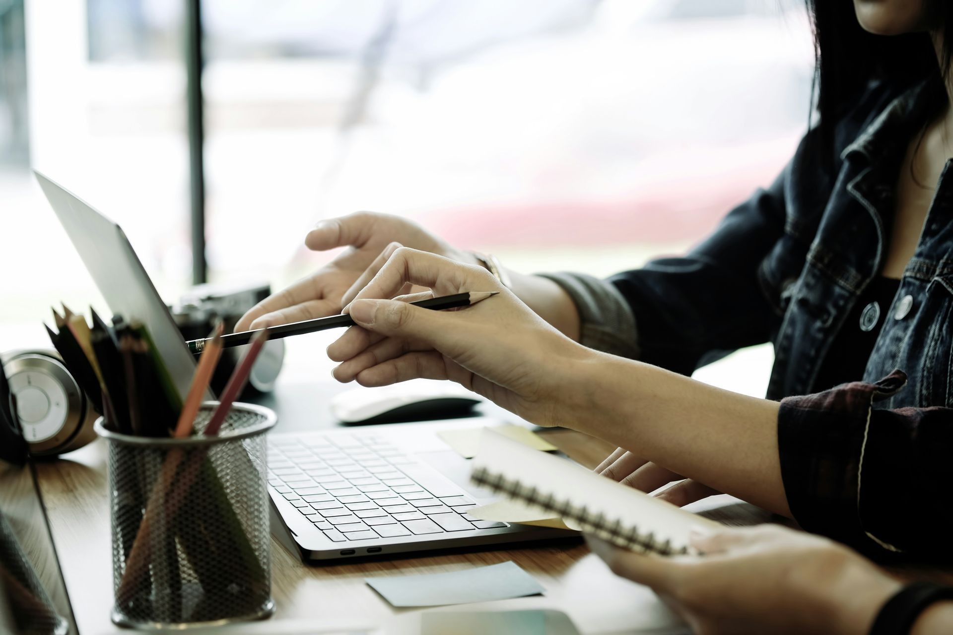 Hands pointing at laptop screen, one holding pencil, other holding notepad, at a wooden desk with colored pencils.