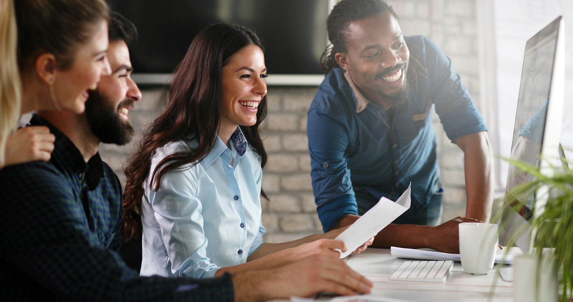 Group of colleagues smiling and looking at a computer screen, possibly during a meeting, in a modern office.