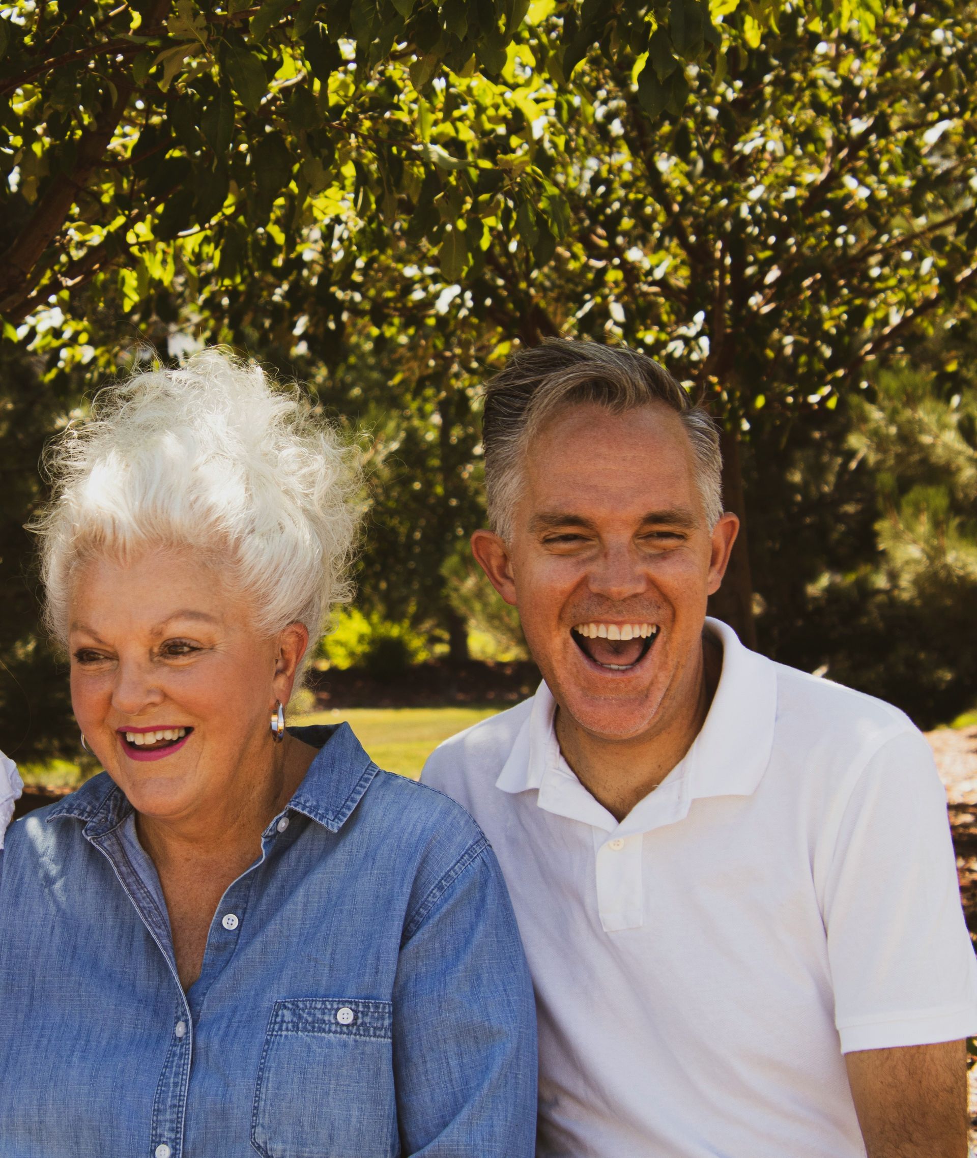 Smiling man and woman outdoors, the man wearing a white shirt, the woman a denim shirt, with leafy trees in the background.
