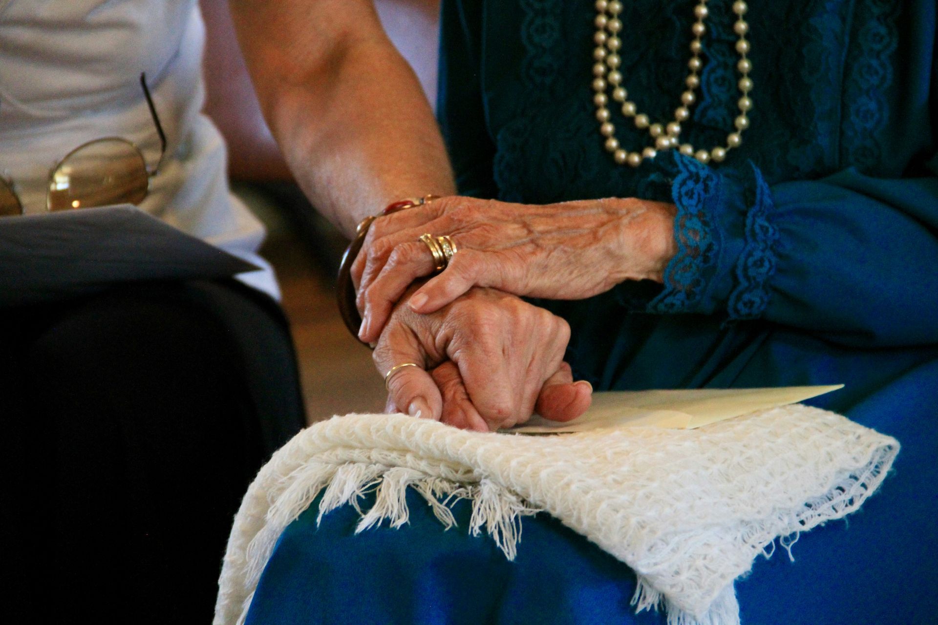 Hands of two people clasped together, one wearing a ring and bracelet, resting on a white fringed cloth.