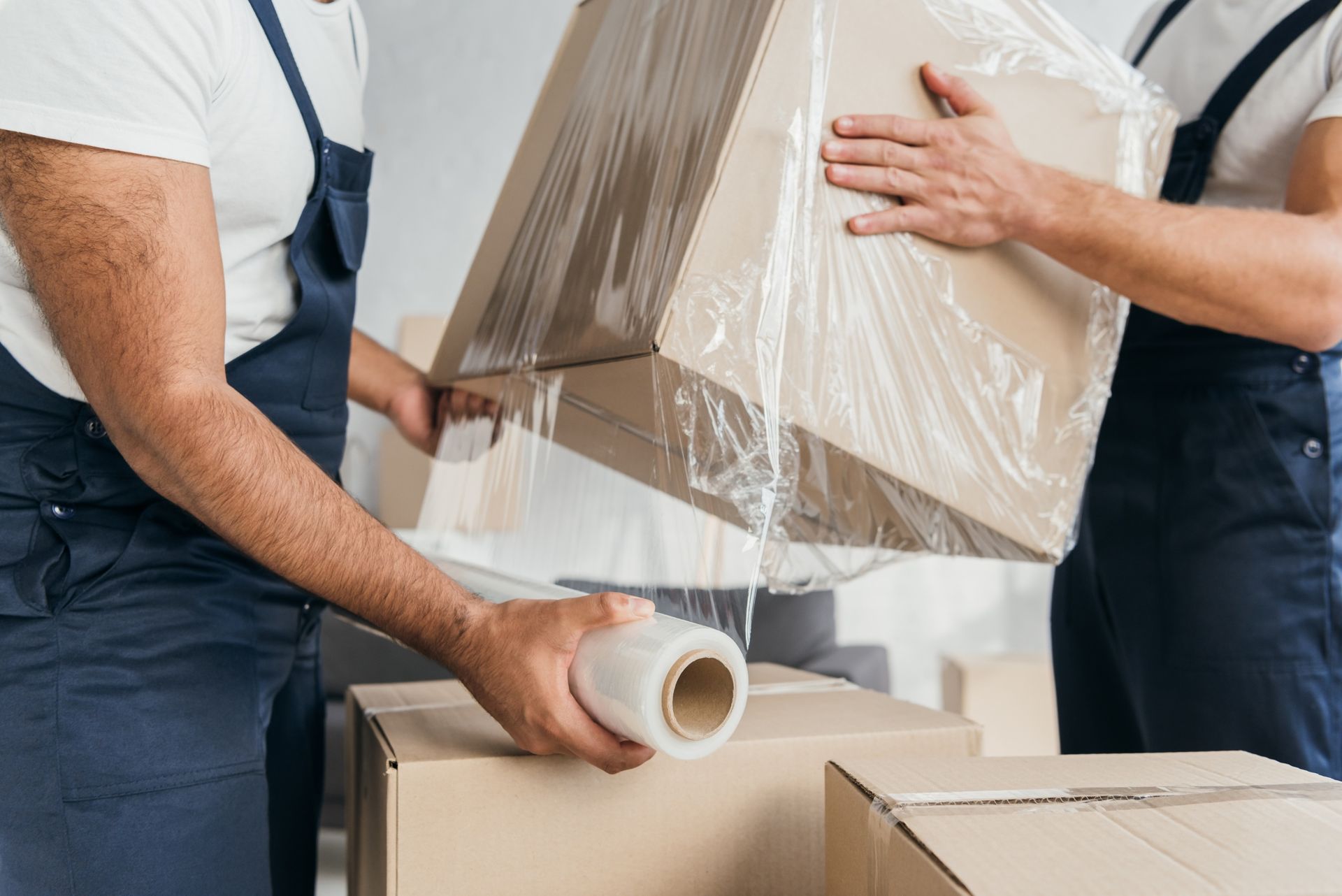 Two movers wrapping a cardboard box in clear plastic beside stacked boxes