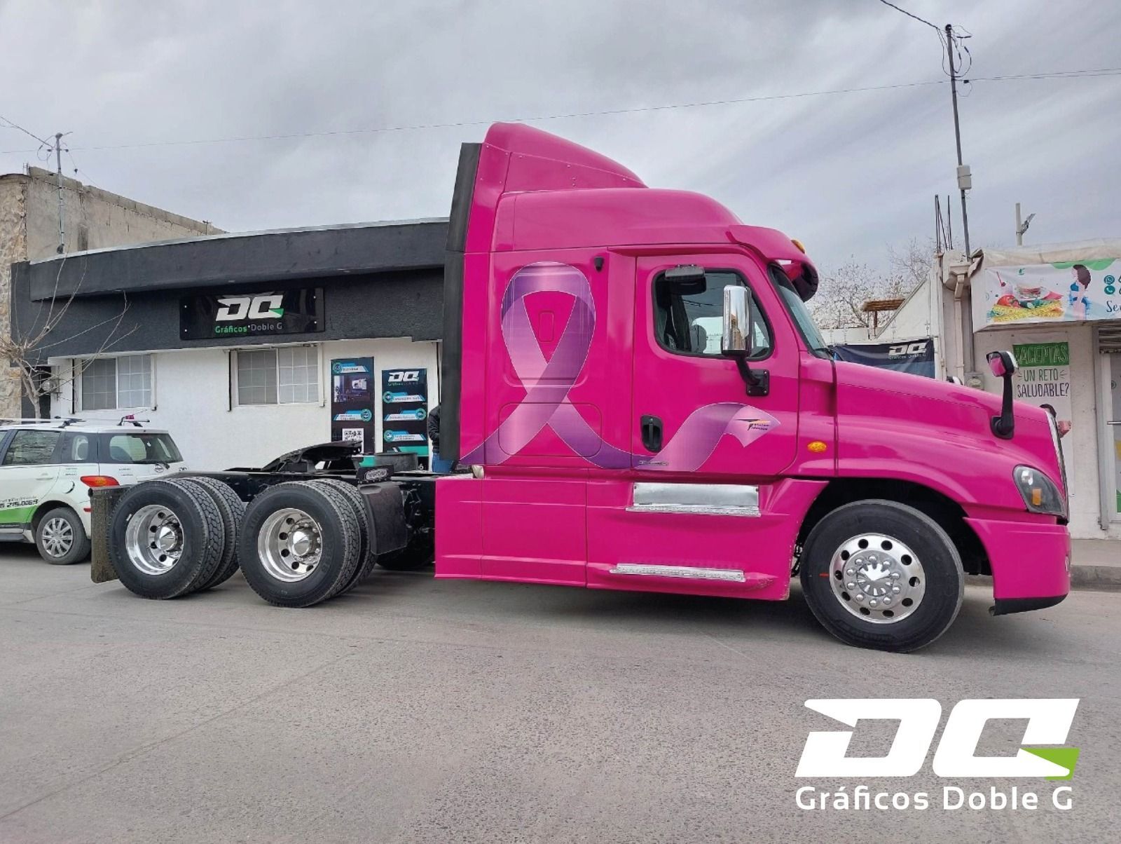Un camión semirremolque rosa con una cinta rosa en el costado está estacionado frente a un edificio.