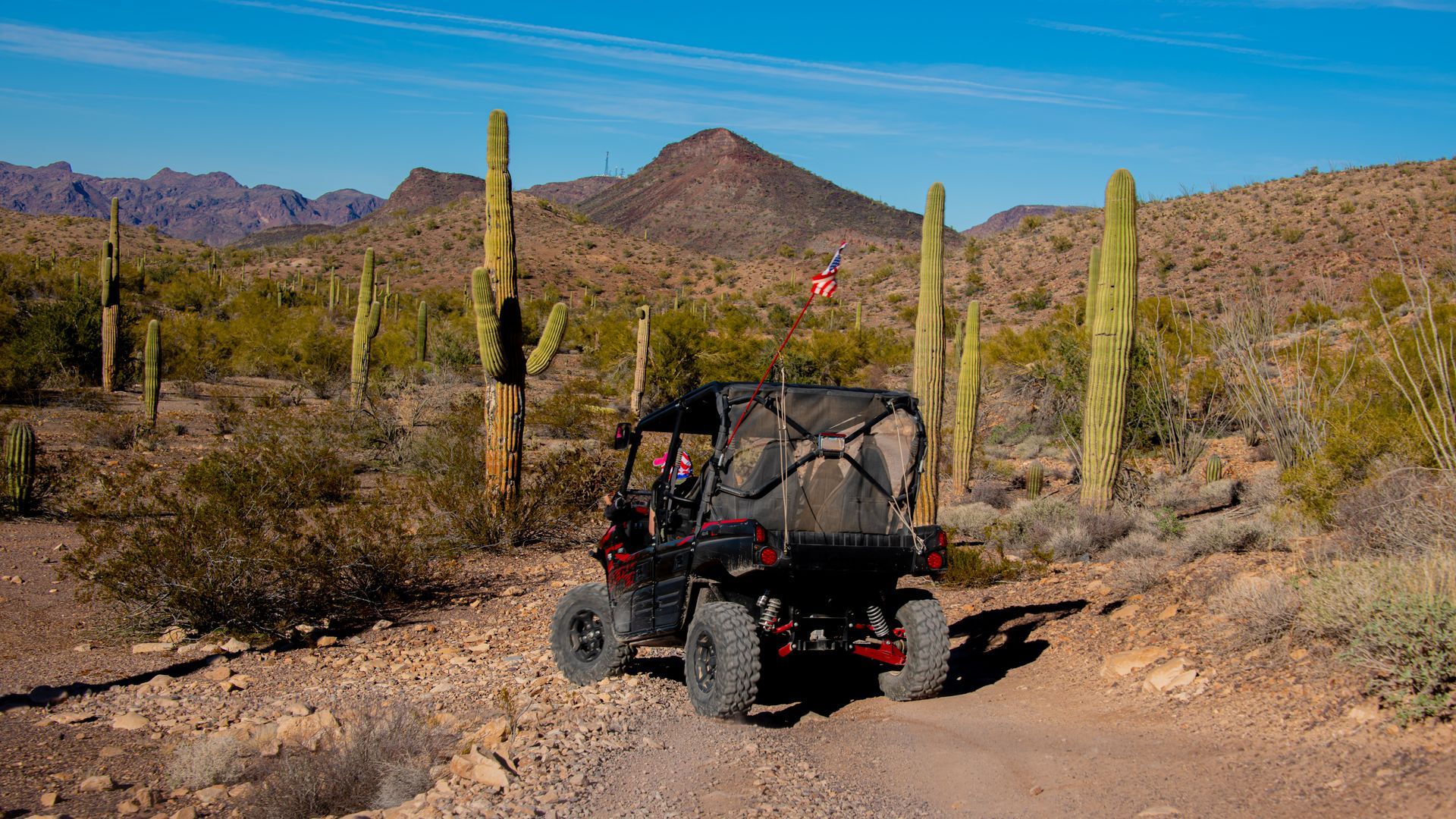 Off-road vehicle driving on a desert dirt road, past cacti, with mountains in the background under a clear blue sky.