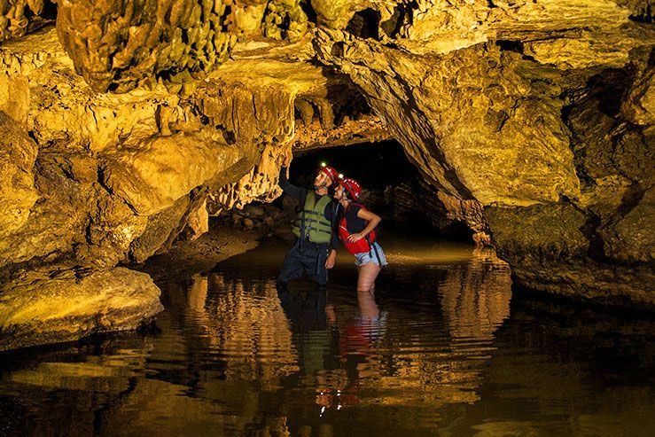 Dos personas de pie en una poza de una cueva iluminada por una luz dorada, reflejadas en el agua