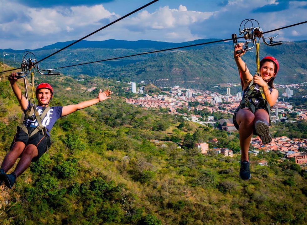 Dos personas en tirolina sobrevolando un valle verde, con una ciudad y montañas al fondo