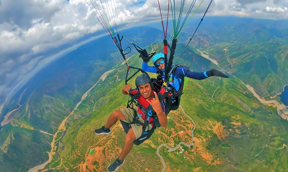 Dos personas vuelan en parapente en tándem sobre montañas verdes bajo un cielo nublado
