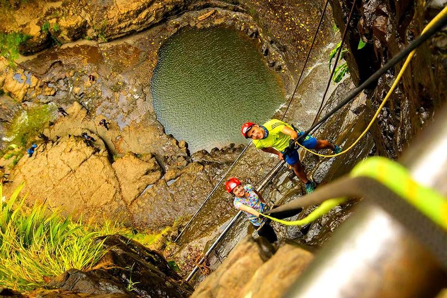 Dos escaladores con casco descienden por un acantilado rocoso junto a una poza circular
