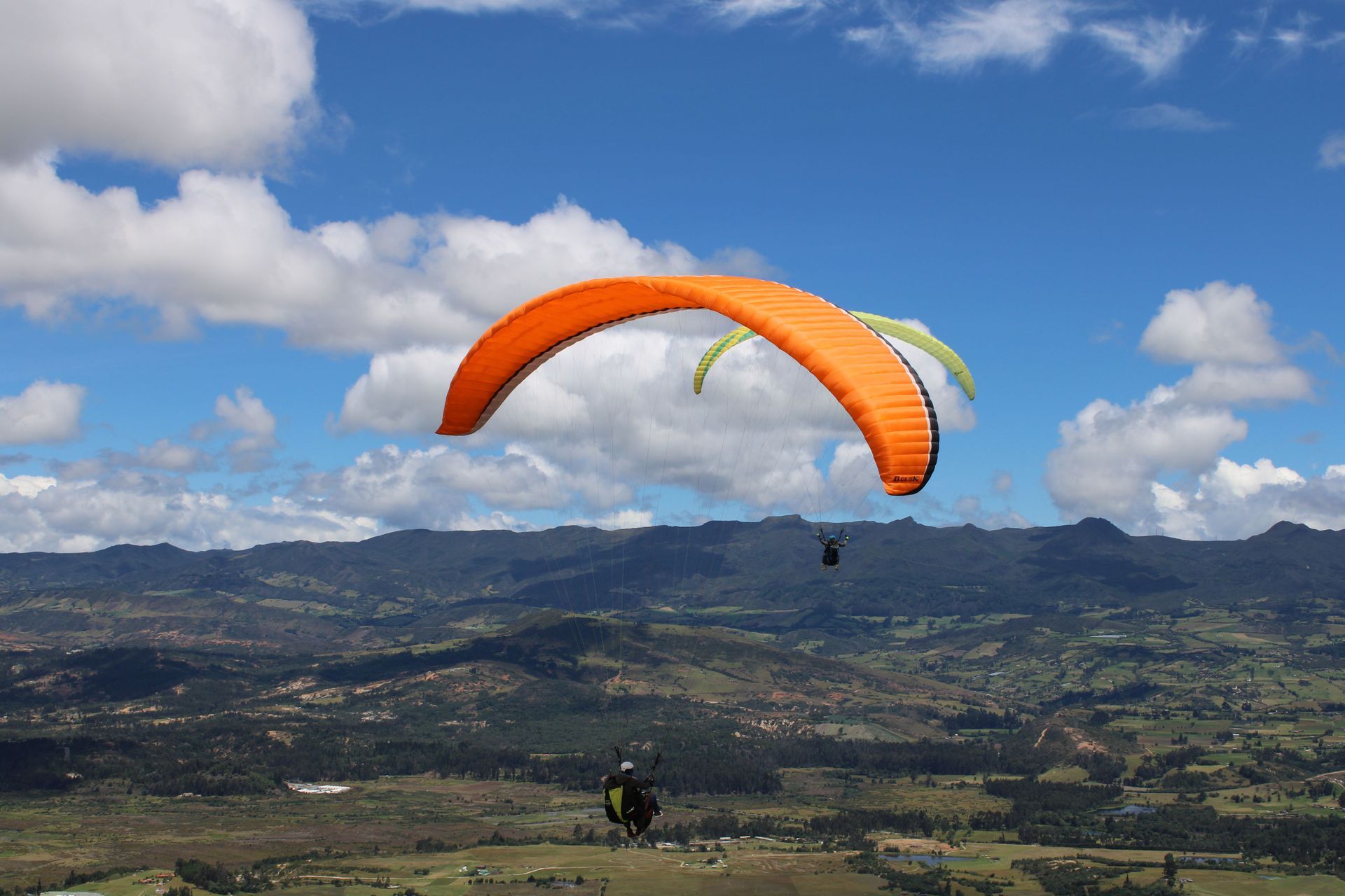 Parapente con una vela naranja volando sobre un valle verde bajo un cielo azul con nubes