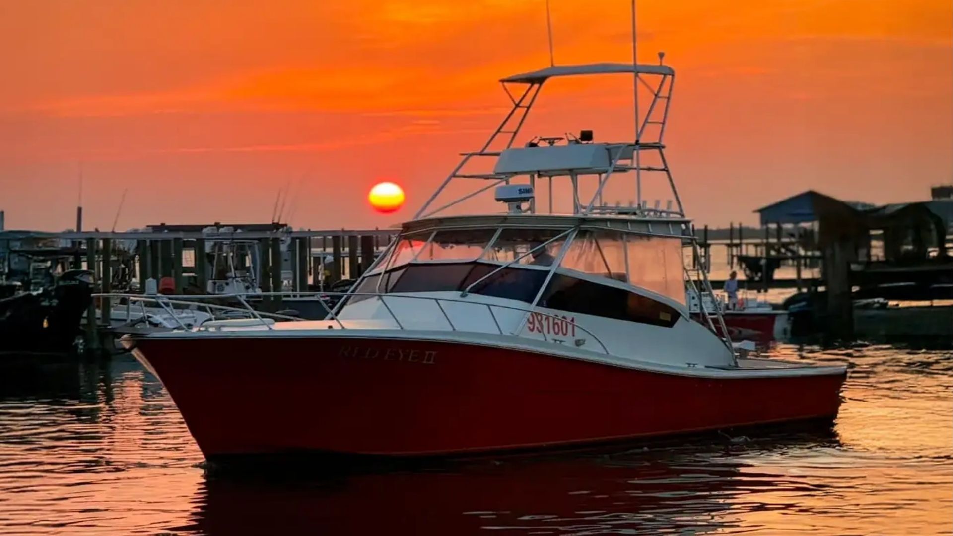 A red and white sport fishing boat docked in a harbor at sunset with a bright orange sun on the horizon.