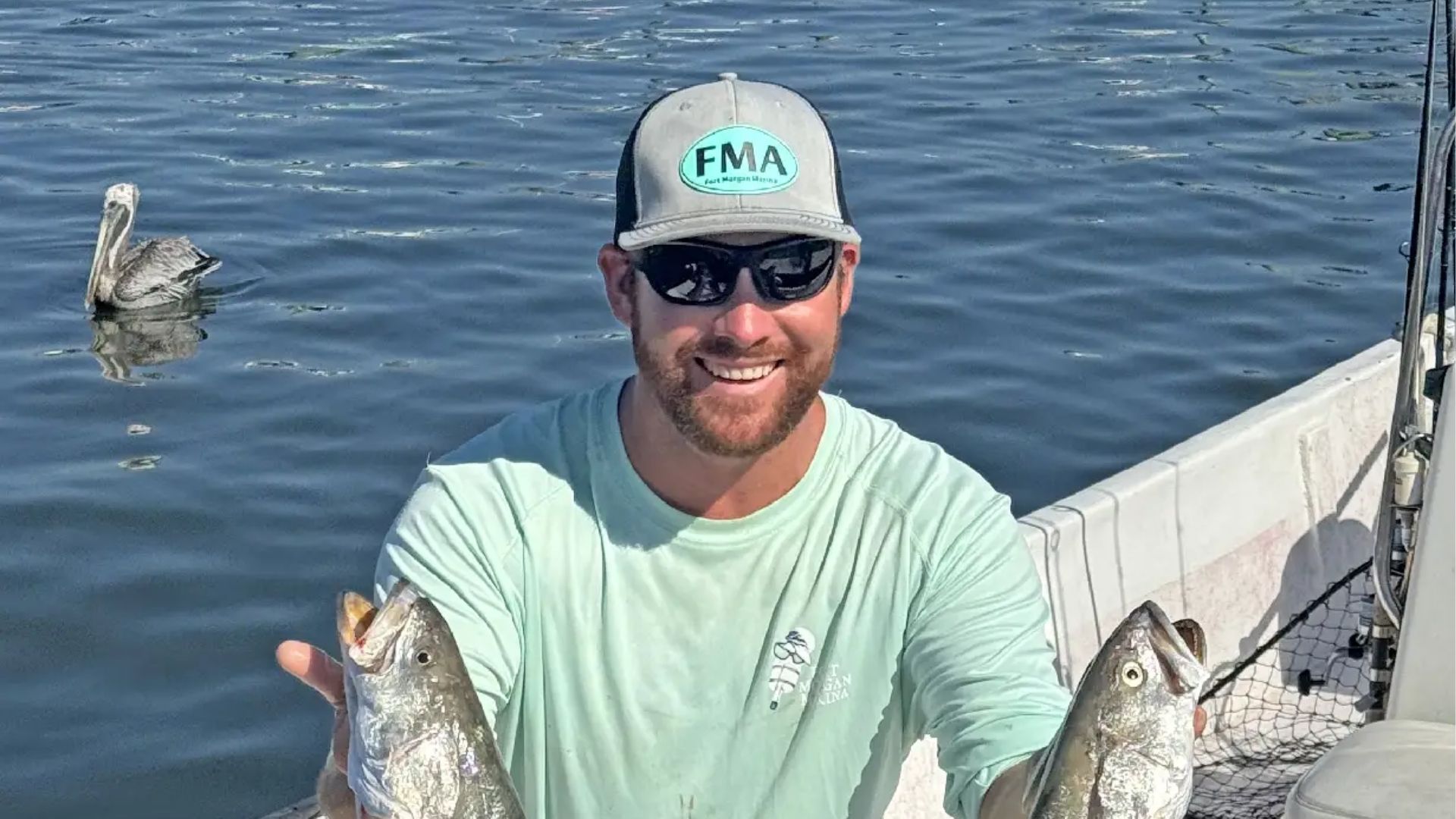 A smiling person wearing a light green shirt and hat holds two fish on a boat, with a pelican swimming in the background.