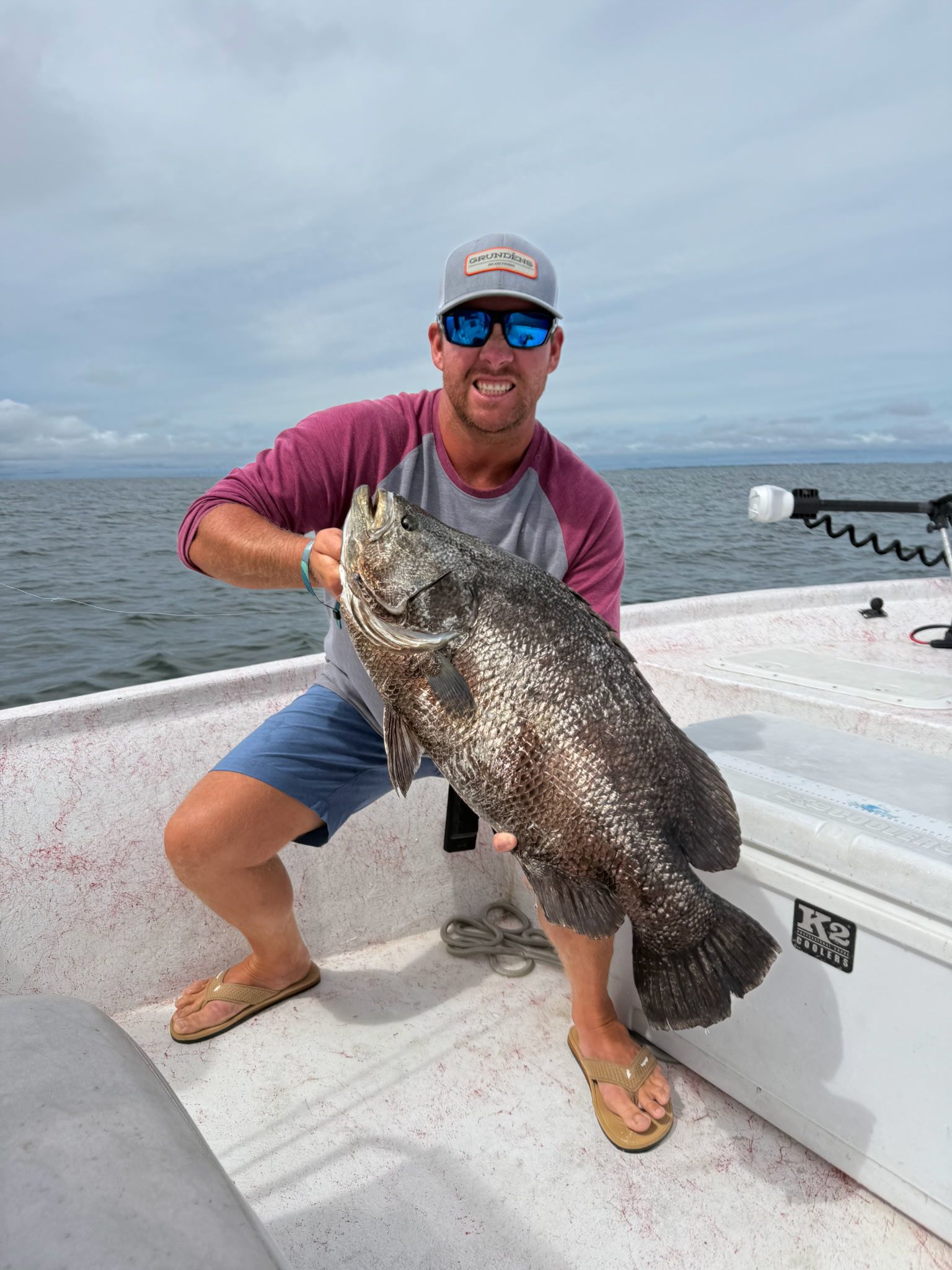 Man smiling on a boat, holding a large speckled fish over the deck with water in the background