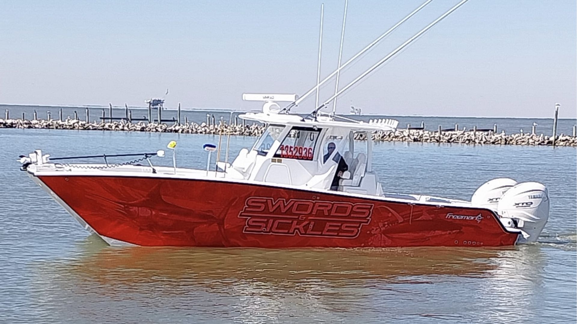 A red and white center console fishing boat named "Swords & Sickles" cruises through calm water near a breakwater.