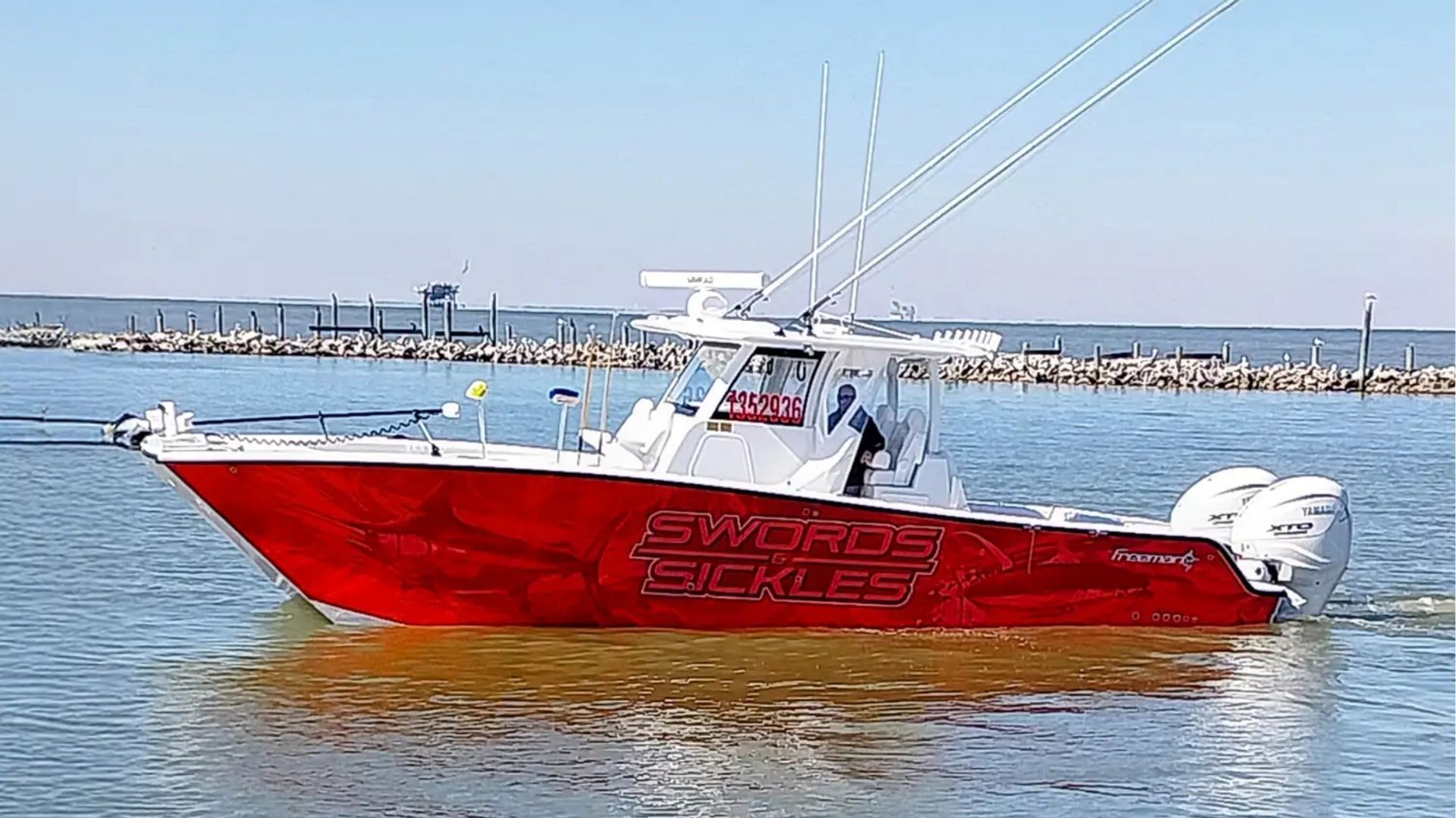 A red center console fishing boat with twin outboard engines cruising on calm water near a rock breakwater.