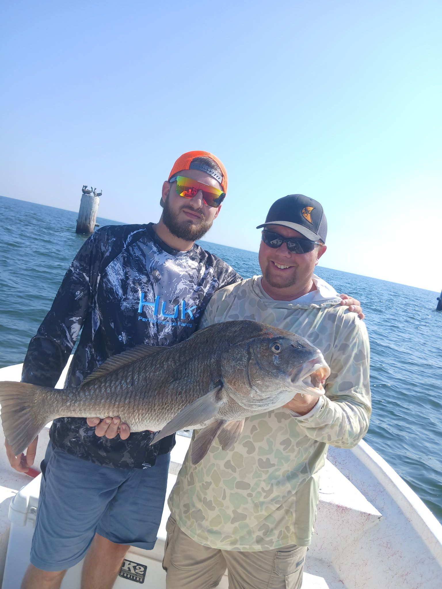 Two men on a boat holding a large fish, with the ocean and blue sky behind them.