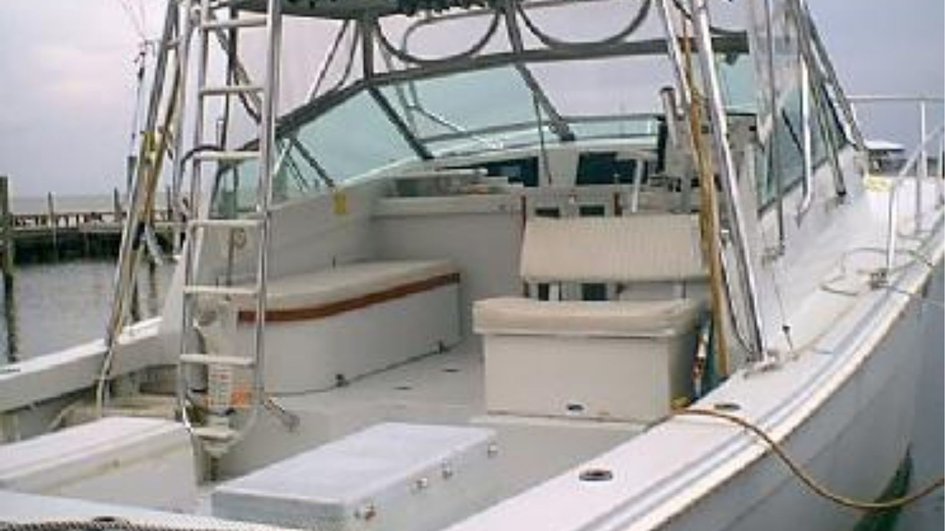 View of the white deck of a sport fishing boat with a ladder, bench seating, and helm chairs at a dock.